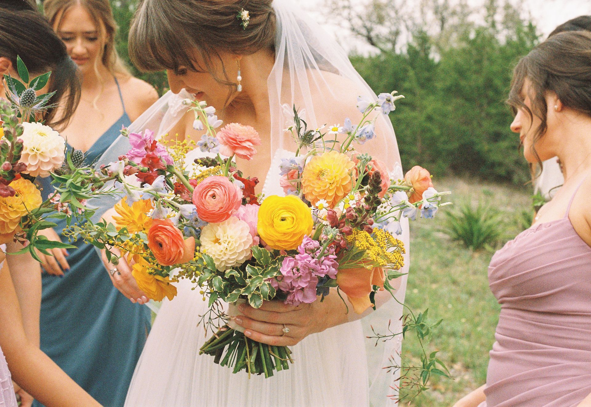 A bride in a white wedding dress holds a large, colorful bouquet of flowers surrounded by bridesmaids in a field.
