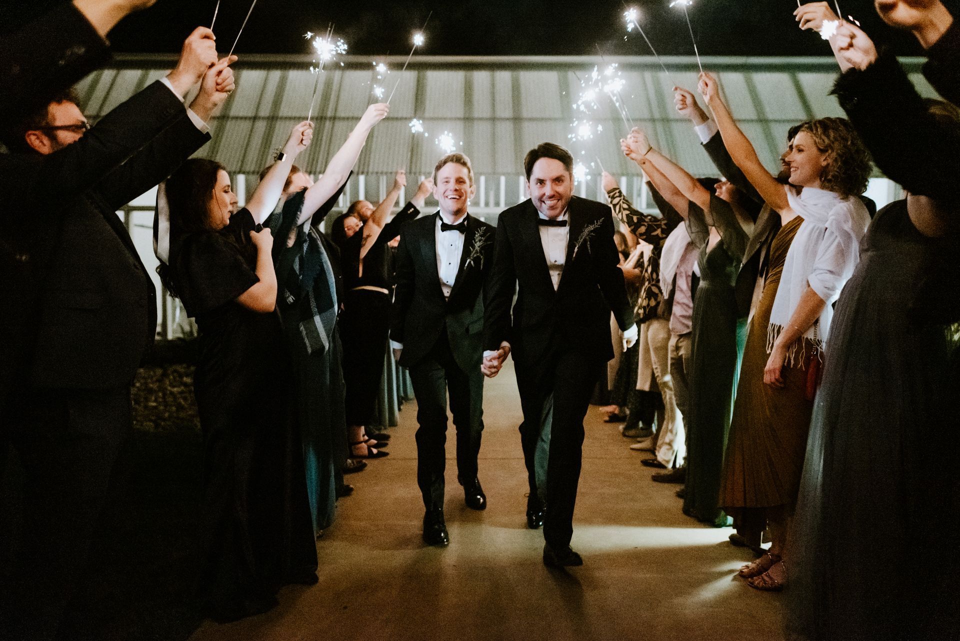 Two men in suits walk down an aisle of wedding guests holding sparklers, smiling as they celebrate.