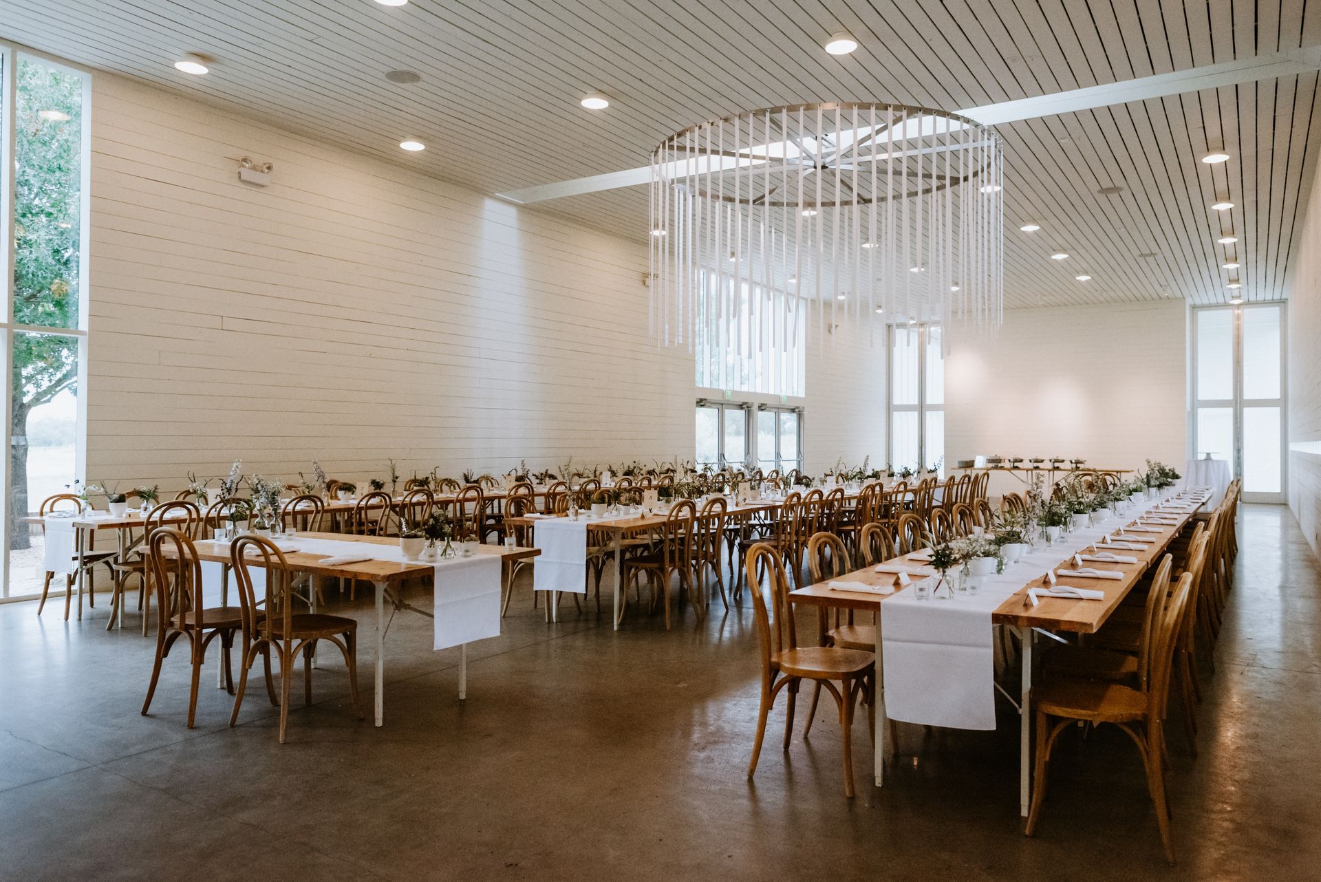 A modern, bright event hall with long, white-covered tables, wooden chairs, and a circular decorative light fixture.