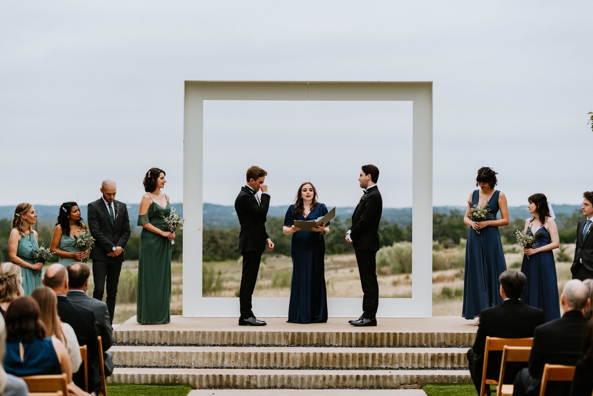 An outdoor wedding ceremony at Prospect House featuring the large white arch frame, featuring a couple, officiant, and guests.