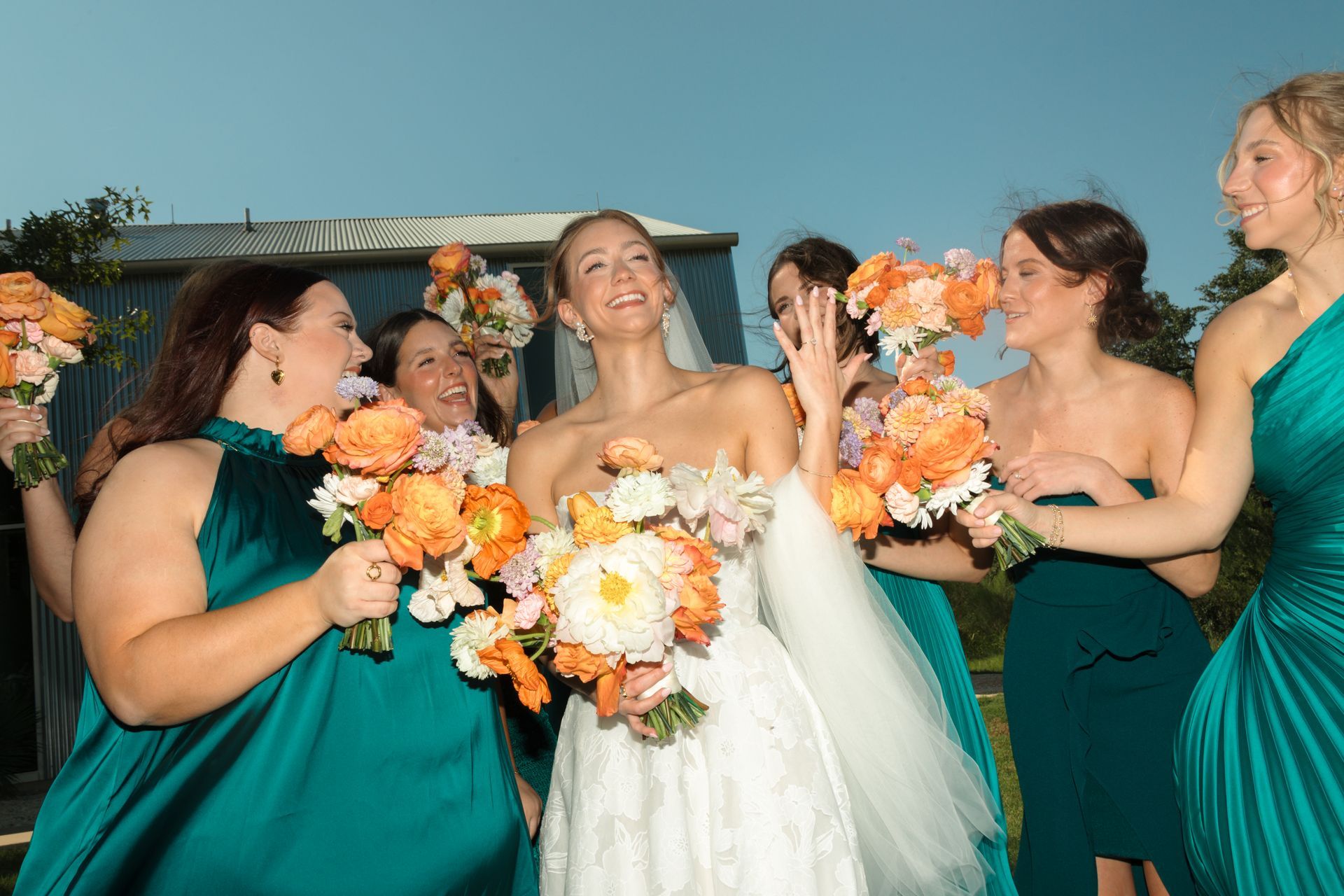A bride in a white gown and four bridesmaids in teal dresses laugh and hold orange bouquets outdoors.