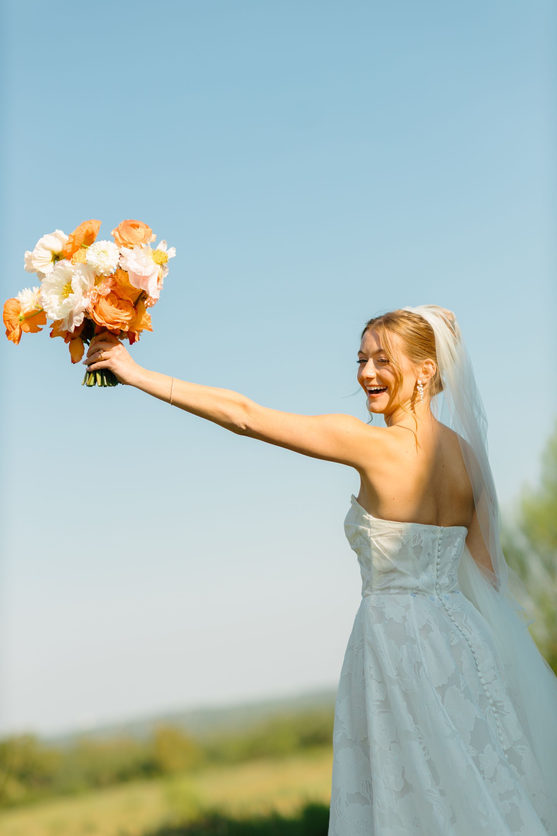 A bride in a strapless white gown and veil holds a bouquet of orange and white flowers up against a clear blue sky.