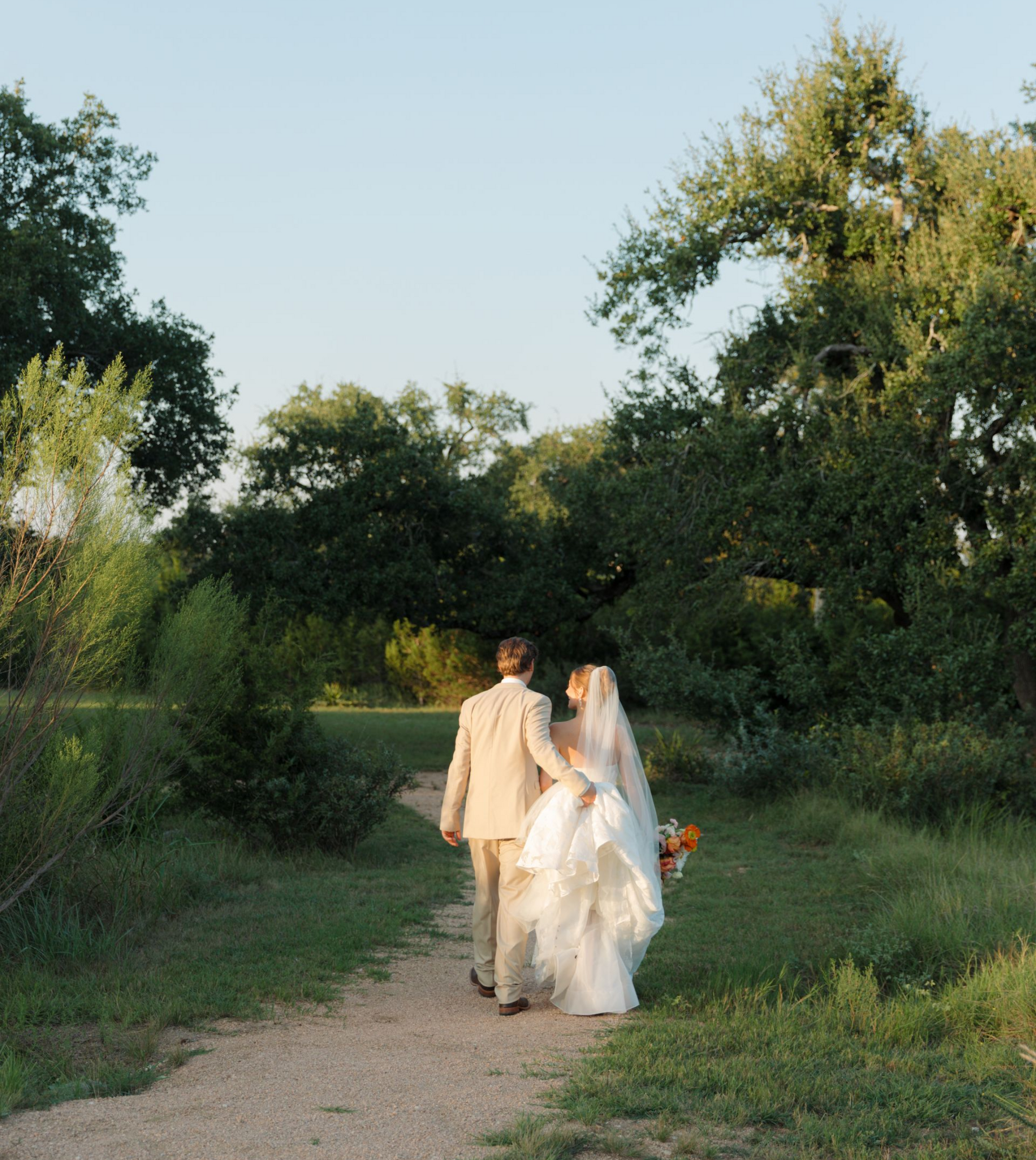 A couple in wedding attire walks away along a dirt path through a grassy, tree-filled landscape in warm light.