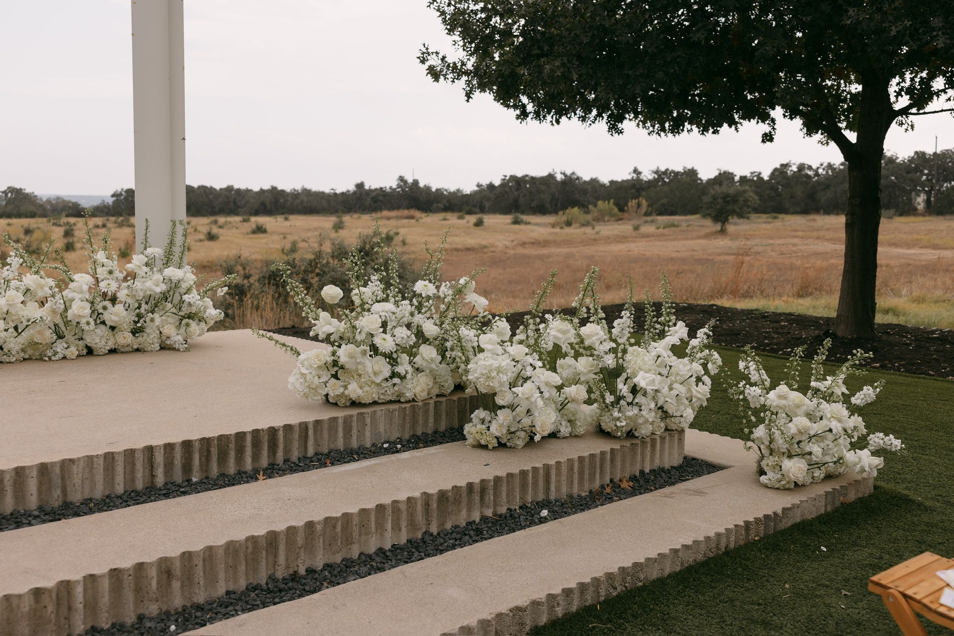White floral arrangements line concrete steps leading to an outdoor landscape with a tree and field in the Texas Hill Country.