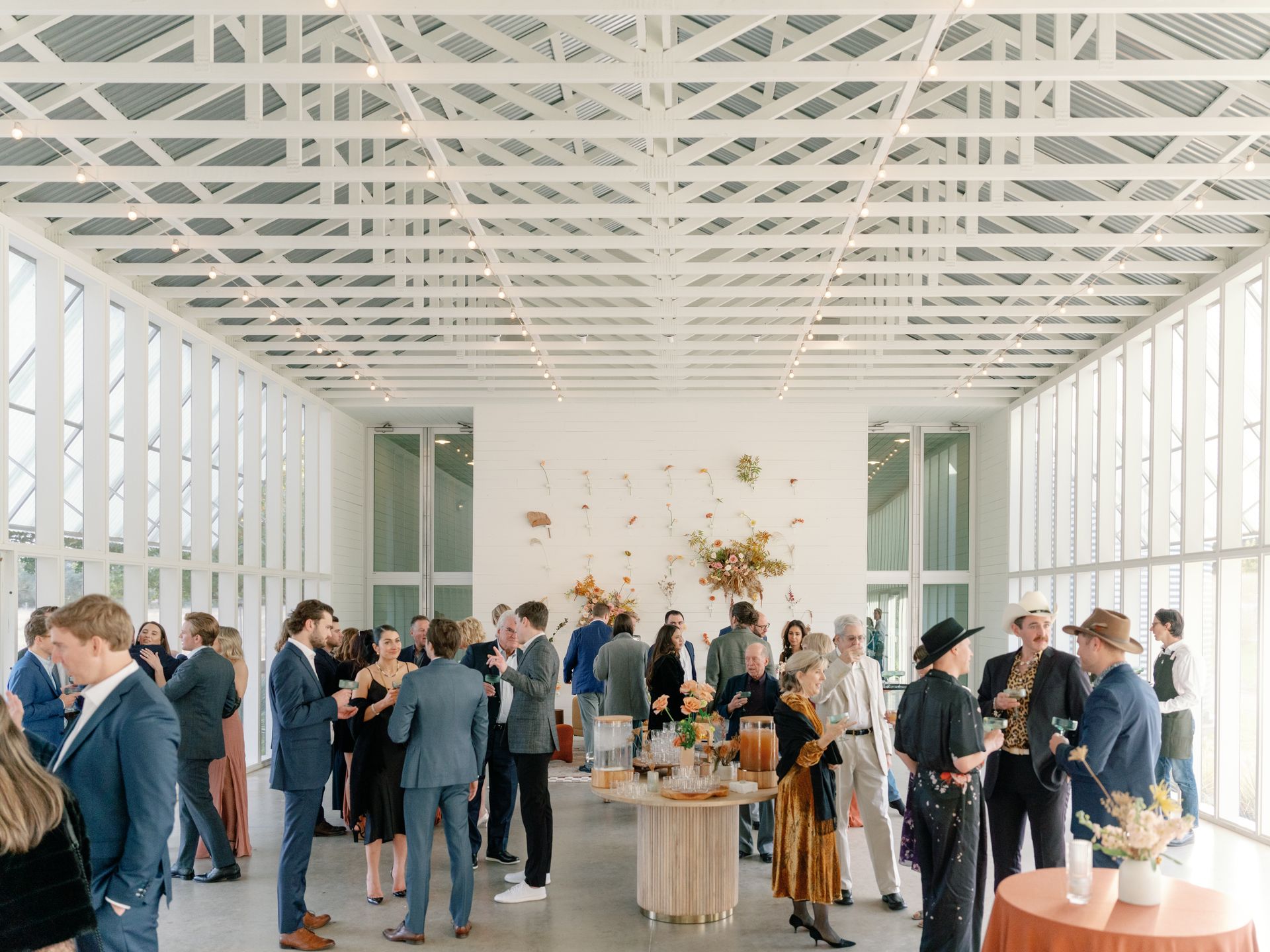 A group of people gathering around tables and mingling in a large room with tall pillared ceilings and beautiful flowers mounted on the plain white wall. 