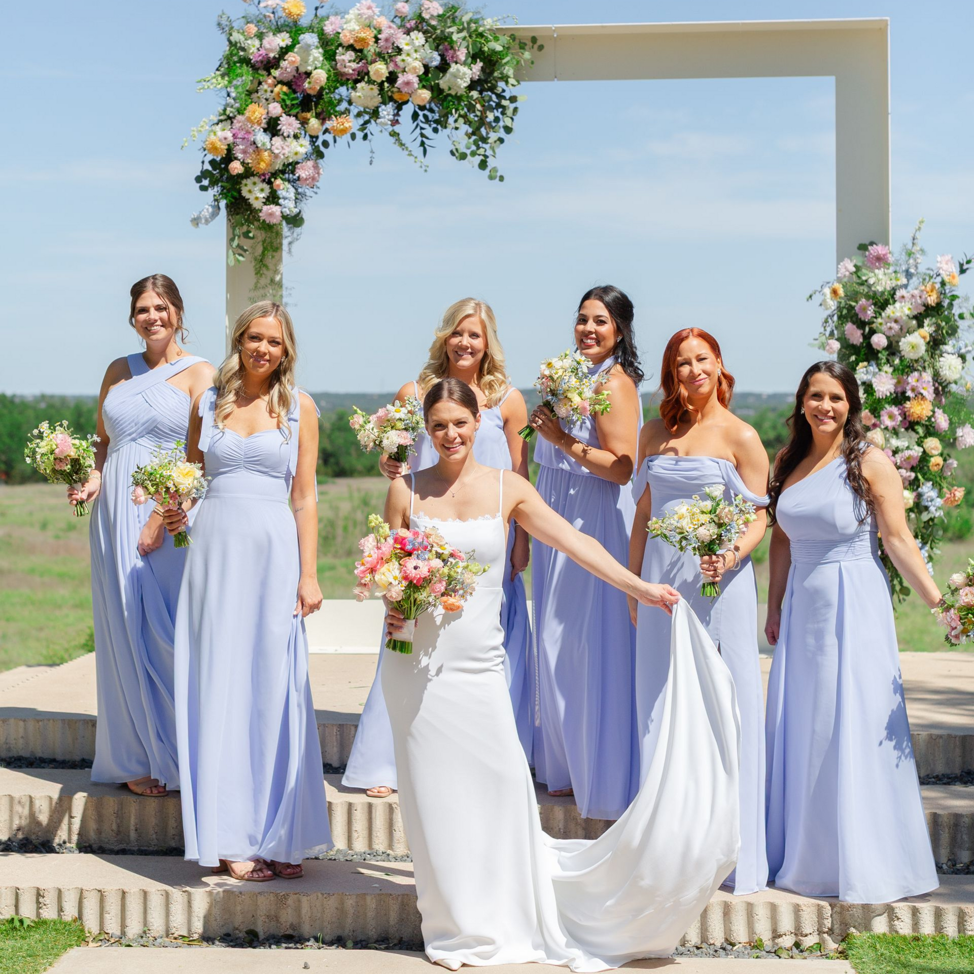 A bride in a white gown and seven bridesmaids in light blue dresses stand outdoors under a floral arch.