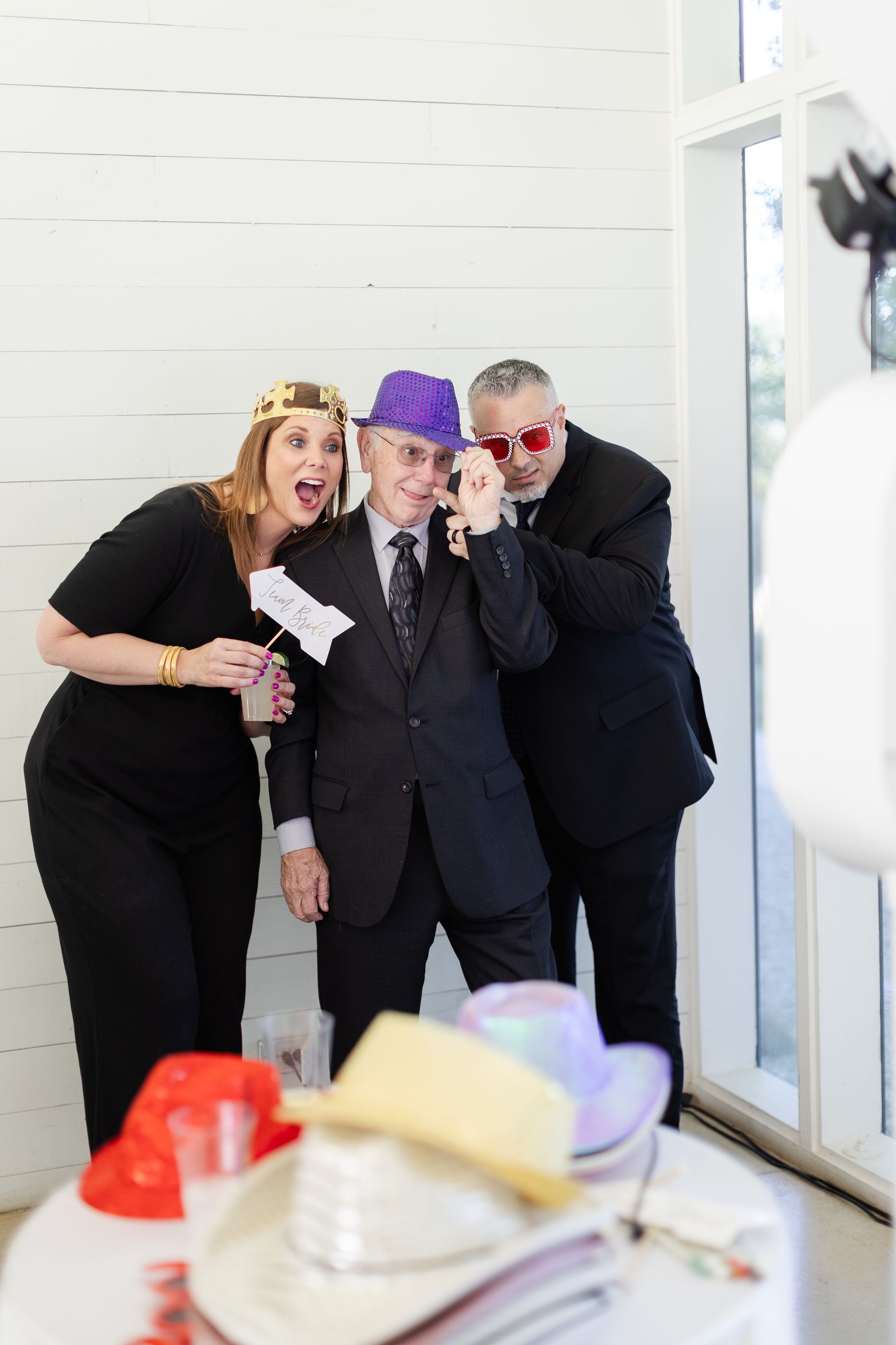 Three people in a photo booth pose with props; one wears a purple hat, one has a crown, and one wears red-tinted glasses.