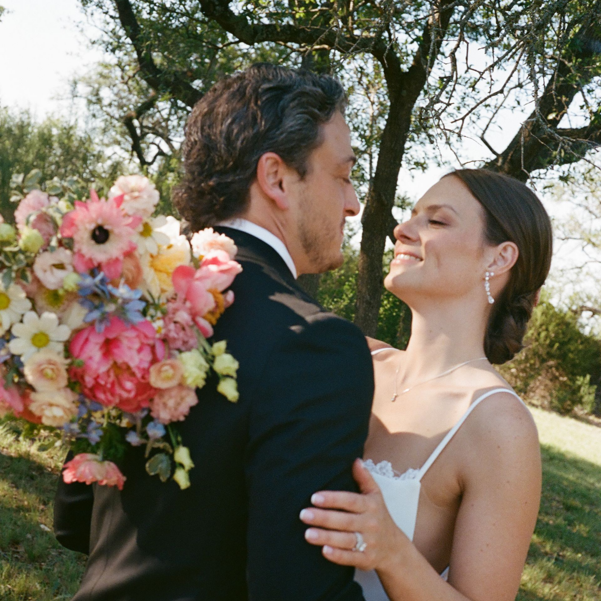 A bride in a white gown and a partner in a dark suit embracing outdoors with a colorful bouquet of flowers.