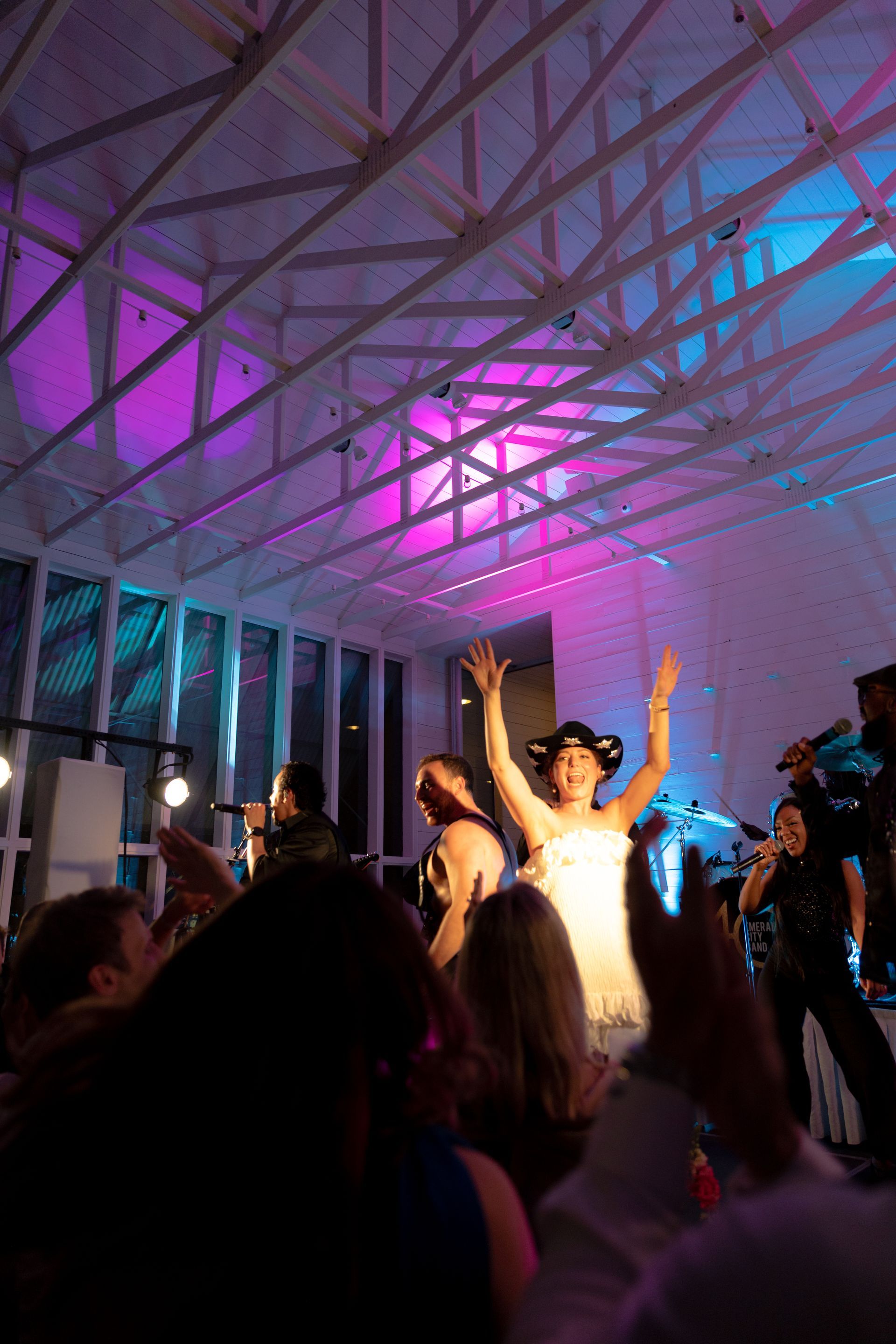 A performer in a white dress and black hat raises both arms on a stage, with band members and an audience in the room.