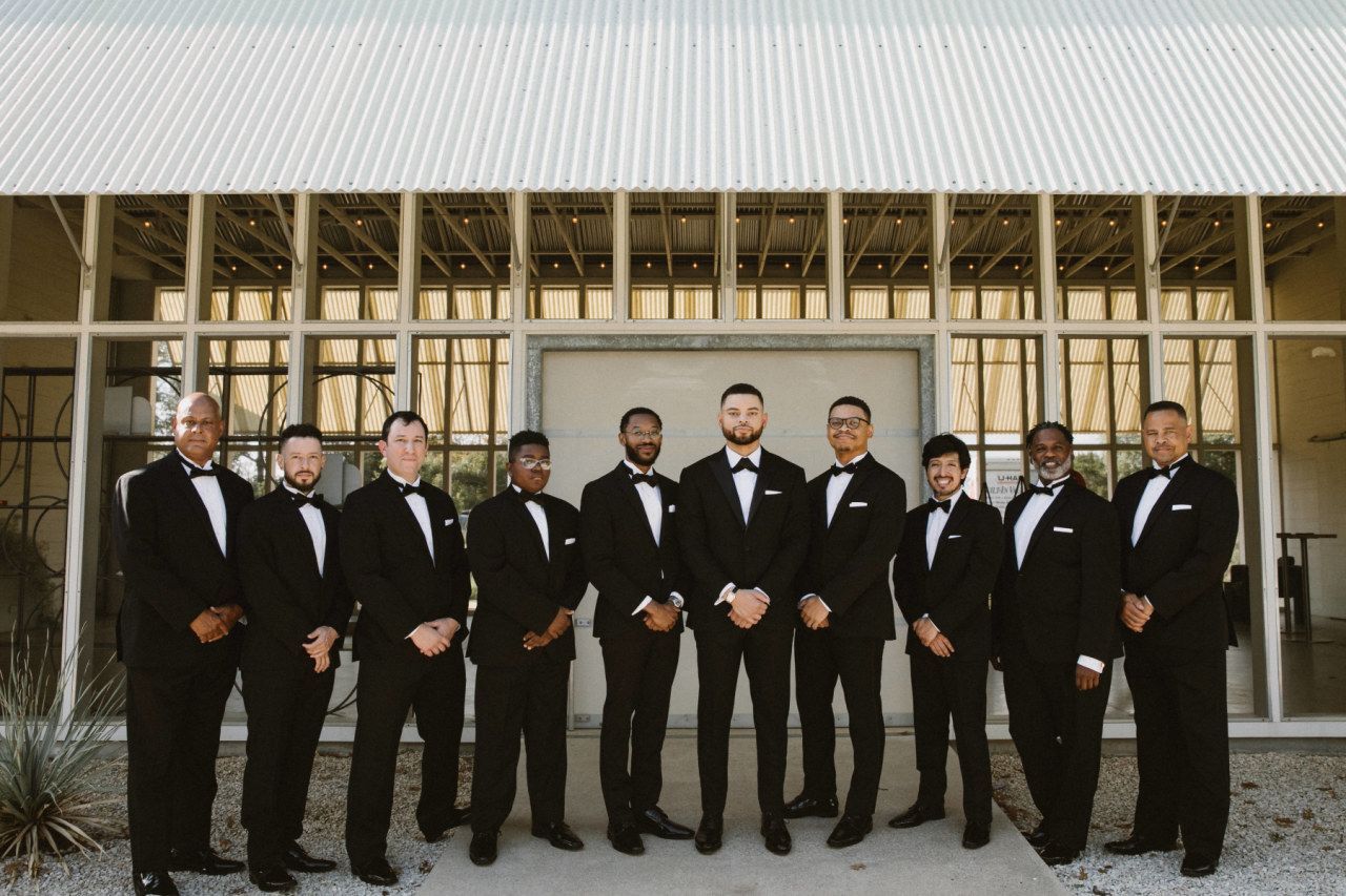 A group of nine men in matching black tuxedos stand in a line before a modern building with large glass windows.