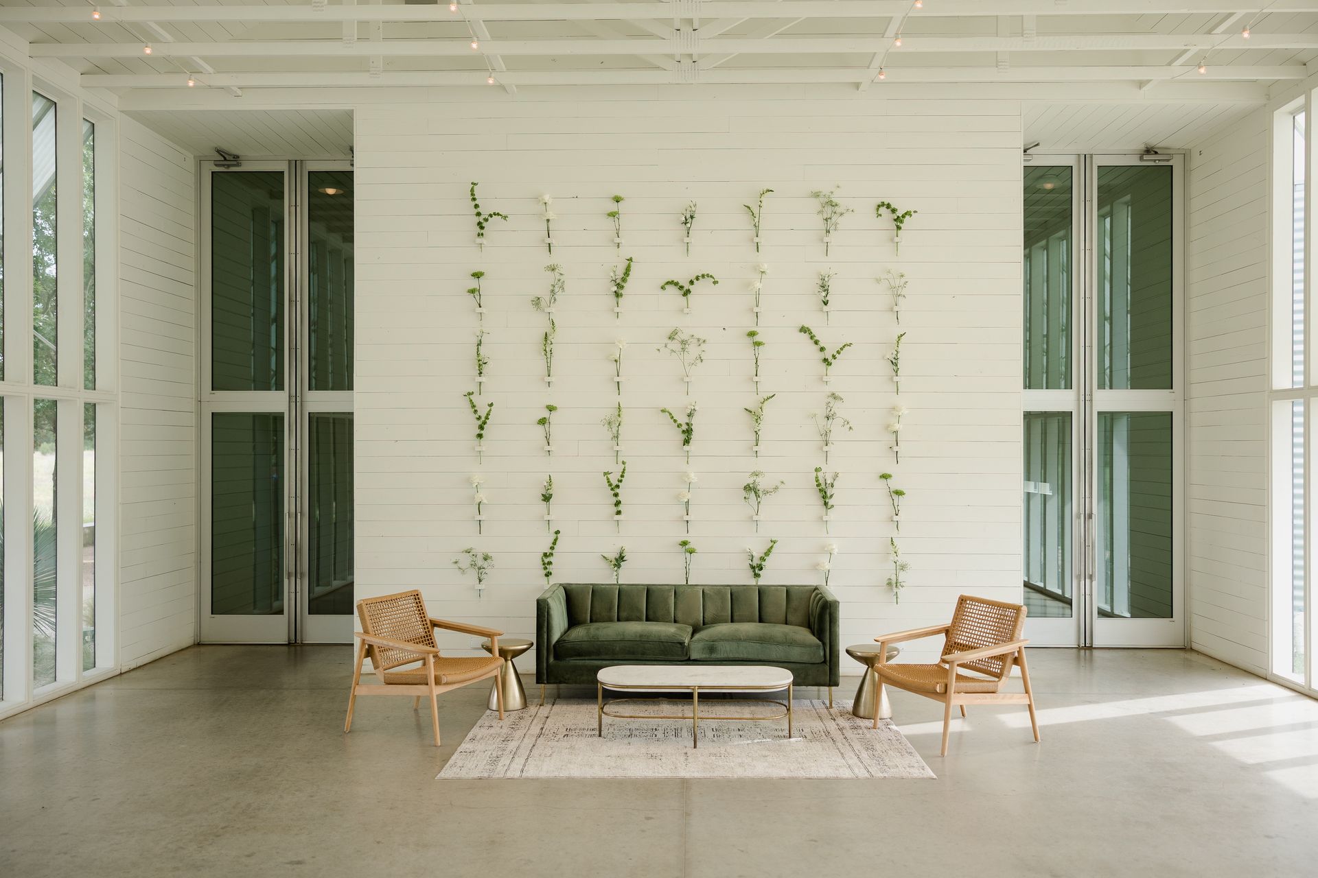 A green velvet sofa and two wooden chairs sit on a patterned rug against a white wall decorated with hanging greenery.