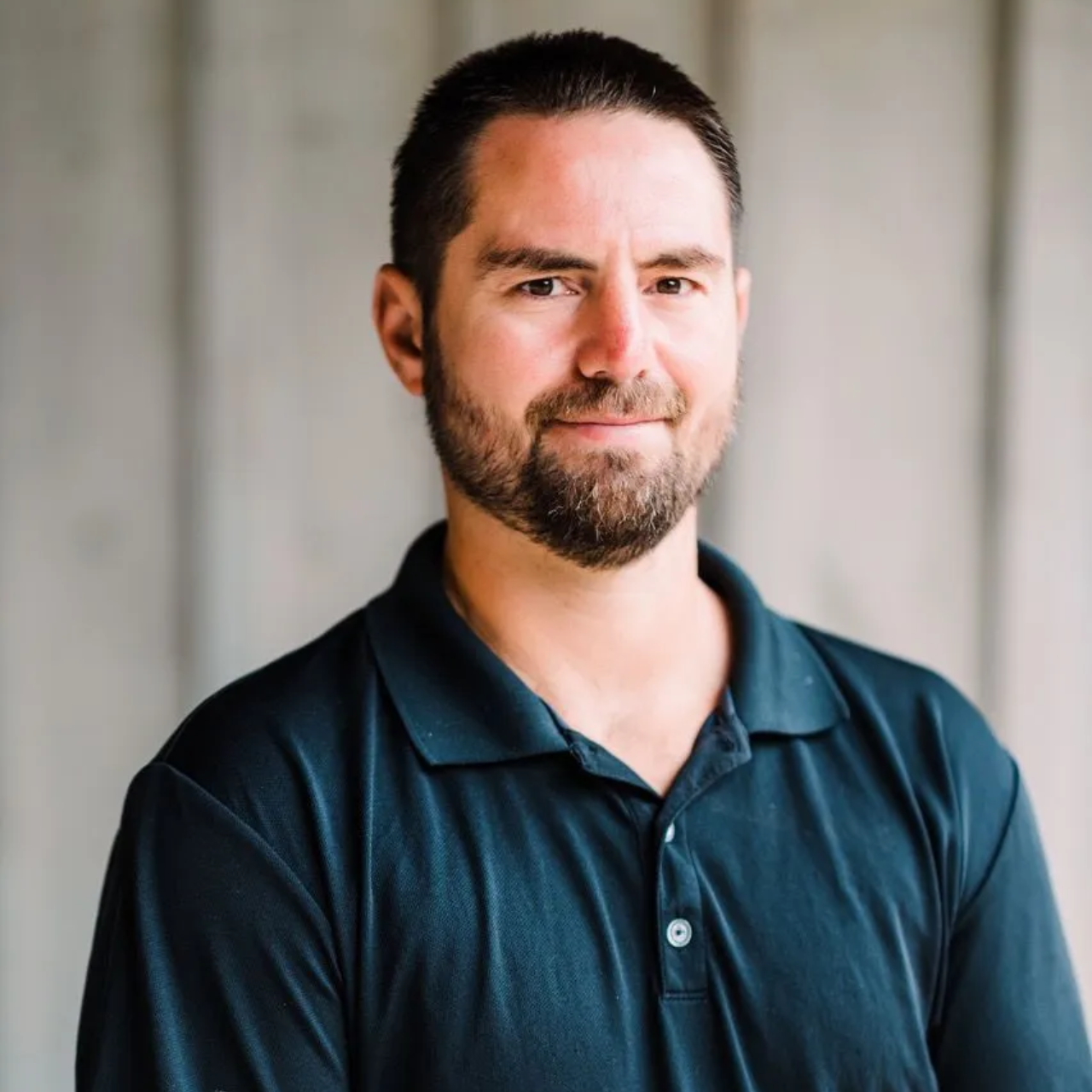 A person with a short beard wearing a dark polo shirt, standing in front of a neutral-toned wooden wall.