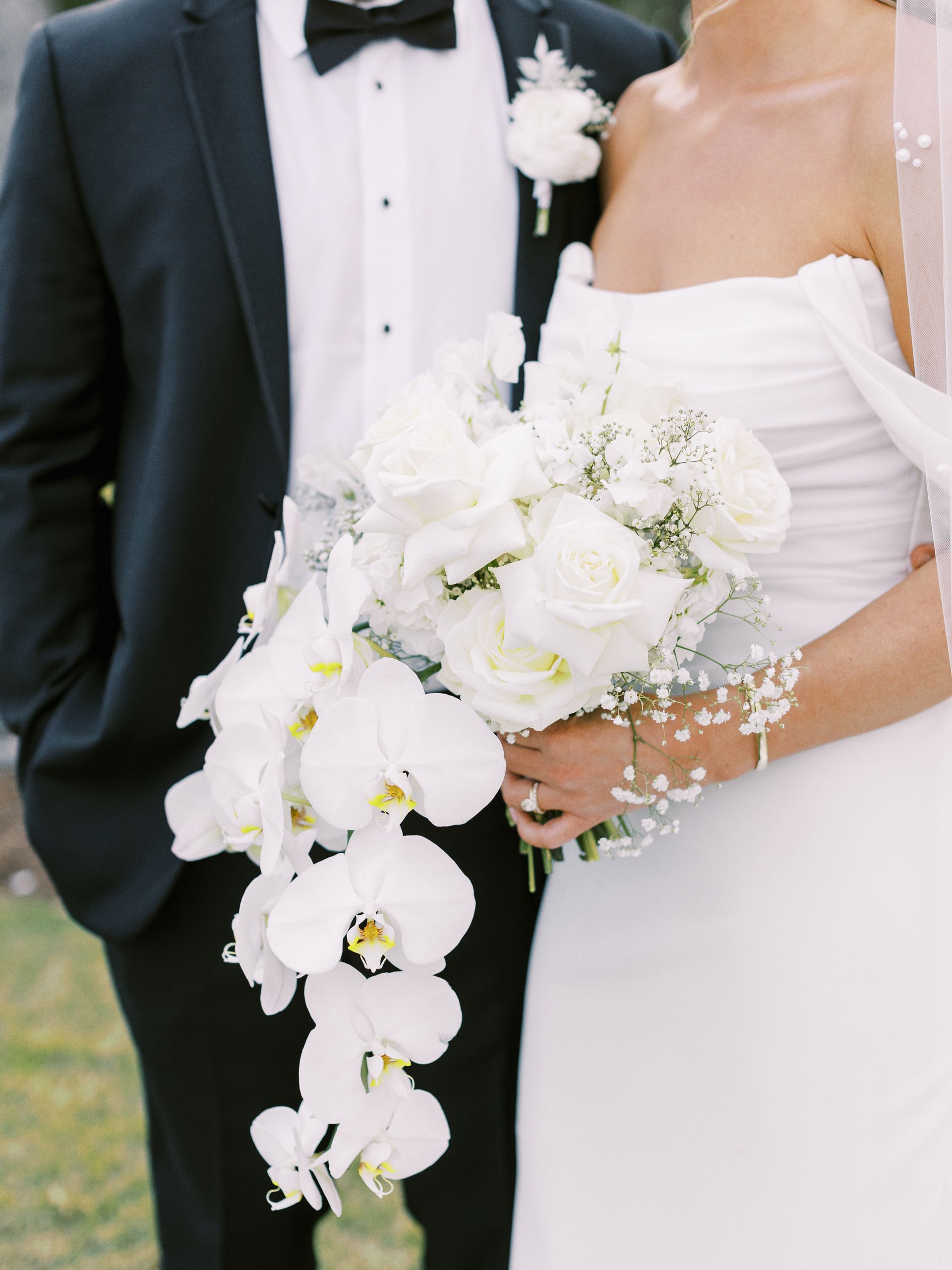A bride in a white wedding gown and a groom in a black suit hold a cascading bouquet of white orchids and roses.