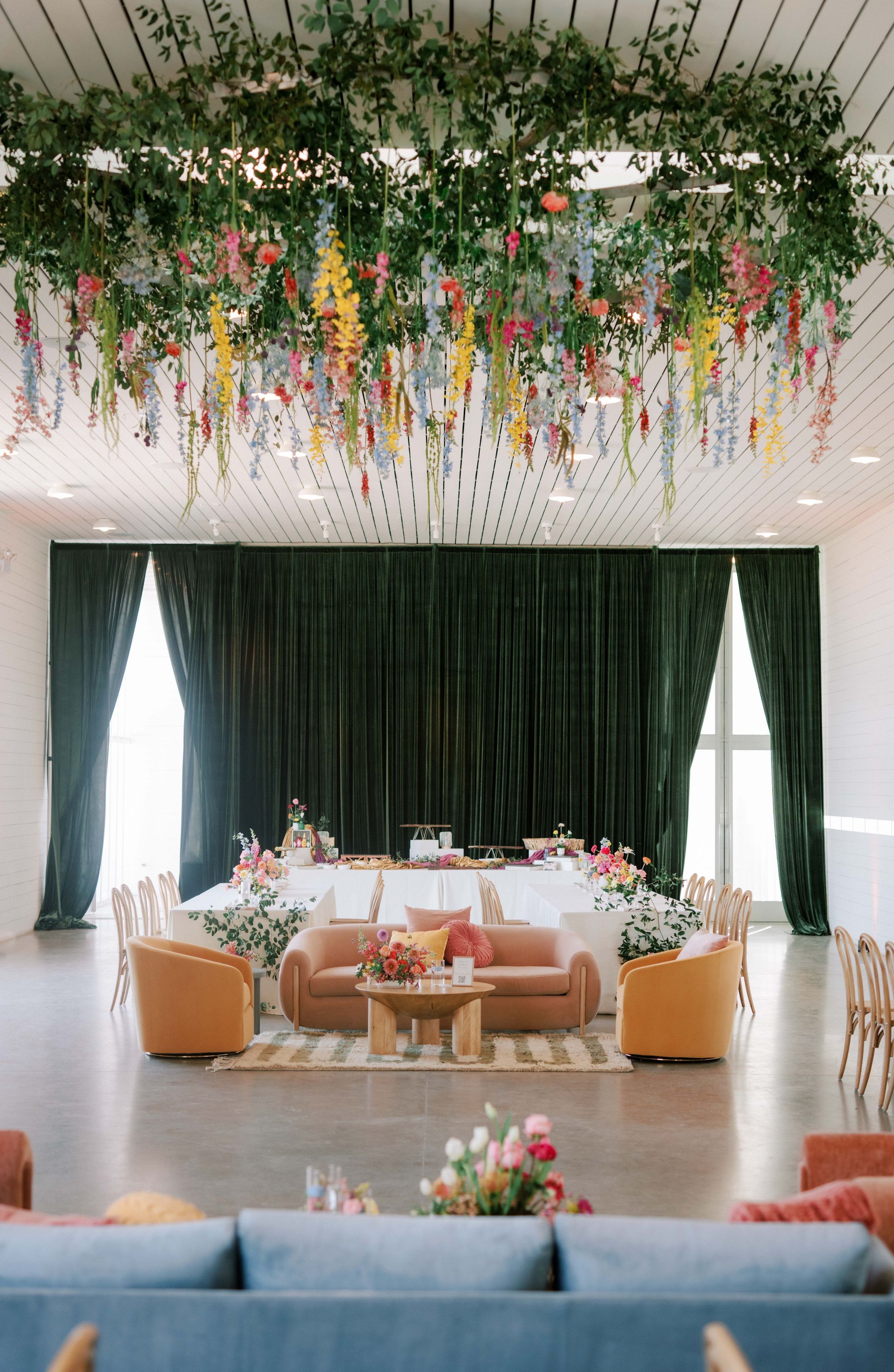 A large room with lounge furniture as well as tables set for eating. There is green drapes as the backdrop of the tables with rainbow flowers hanging from the ceiling.
