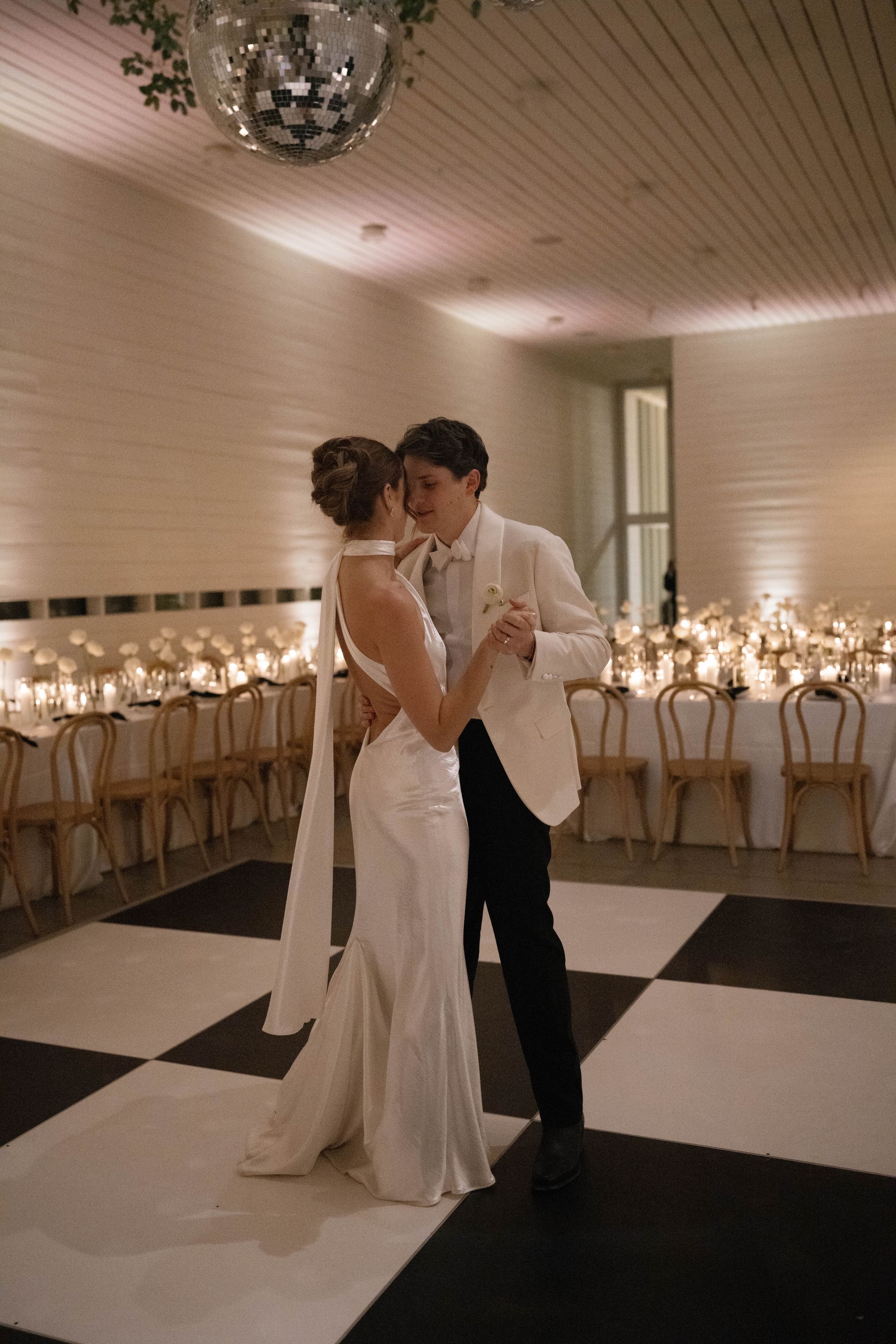 A couple dances on a black-and-white checkered dance floor in a room with warm lighting, candles, and a disco ball.