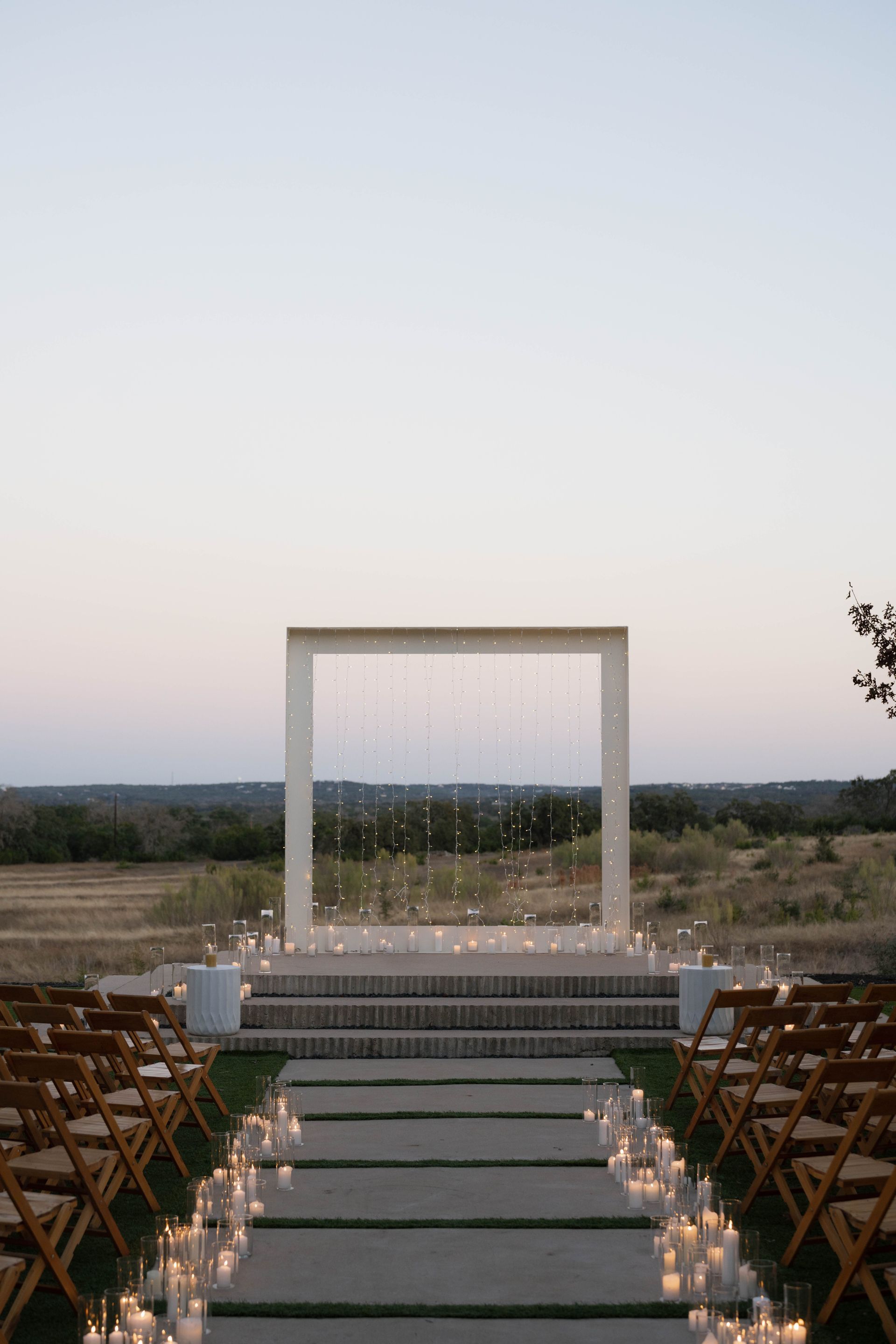 A wedding ceremony setup with rows of wooden chairs, a stone aisle lined with candles, and a large, square white arch.