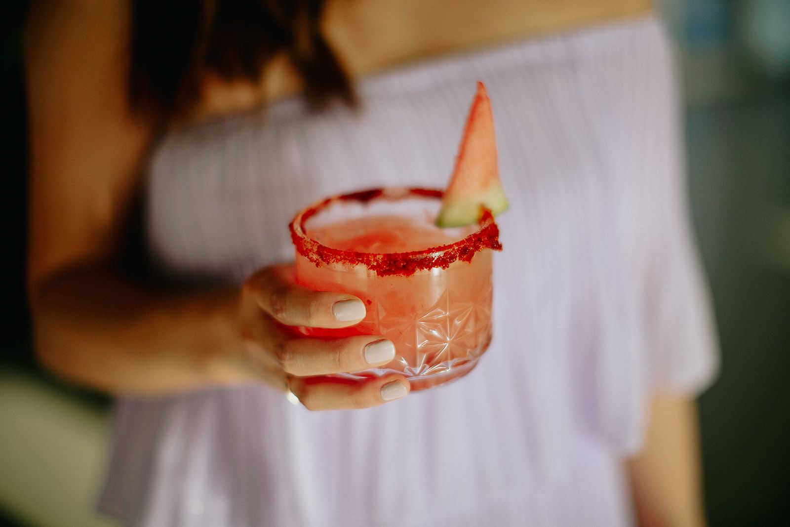 A wedding guest holds a cocktail in a textured glass with a red-rimmed edge and a watermelon slice garnish.