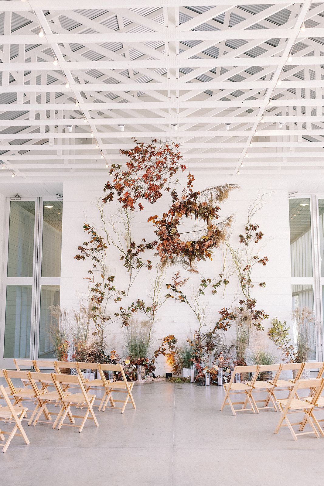Wedding ceremony setup with wooden folding chairs facing a floral wall installation in a bright, modern indoor space.