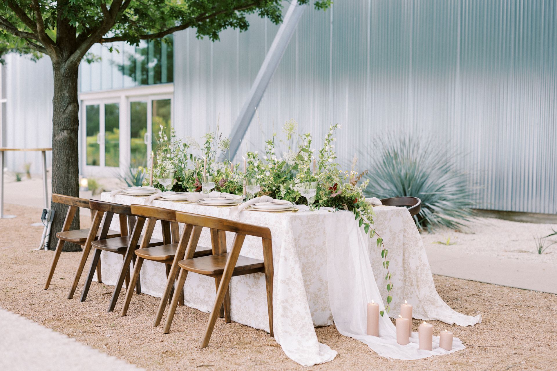 A long table with a white tablecloth and green and white florals cascading down the middle with wooden chairs around in front of a building.