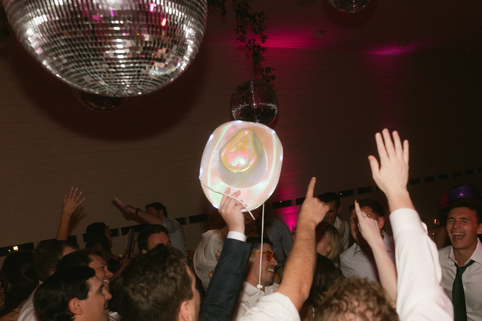 A festive crowd celebrates at a party under disco balls, with one person holding a cowboy hat high in the air.