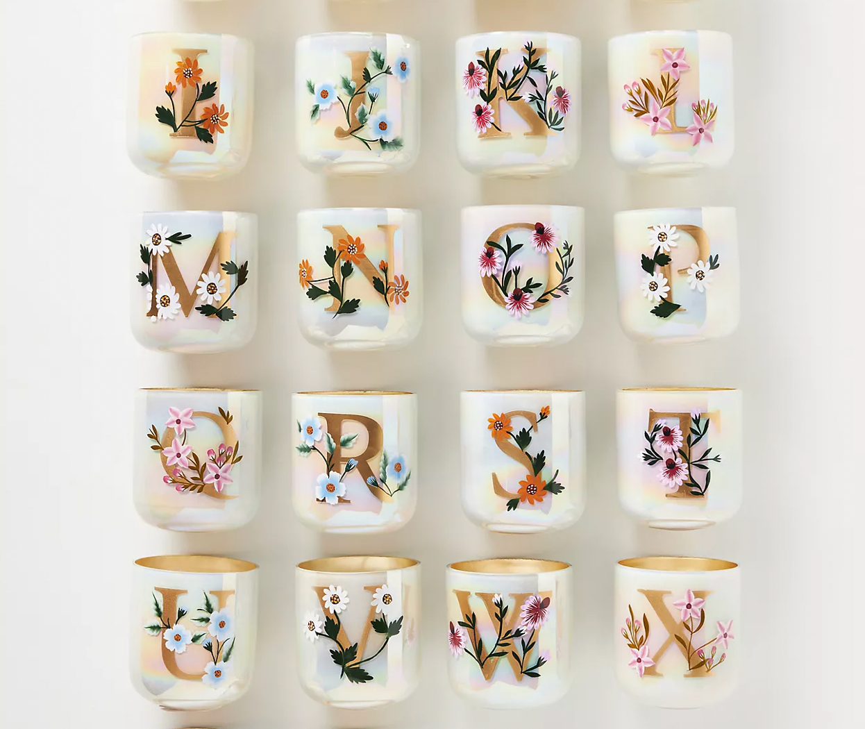 A grid of 16 white mugs, each featuring a large gold letter decorated with colorful floral illustrations.