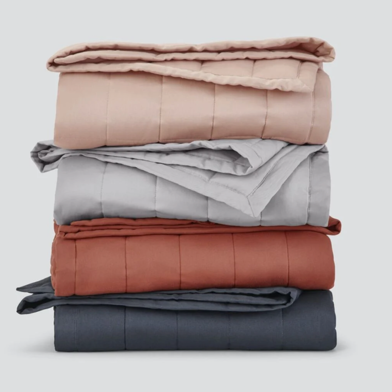 A stack of four folded, quilted blankets in shades of blush pink, light gray, terracotta, and charcoal gray.