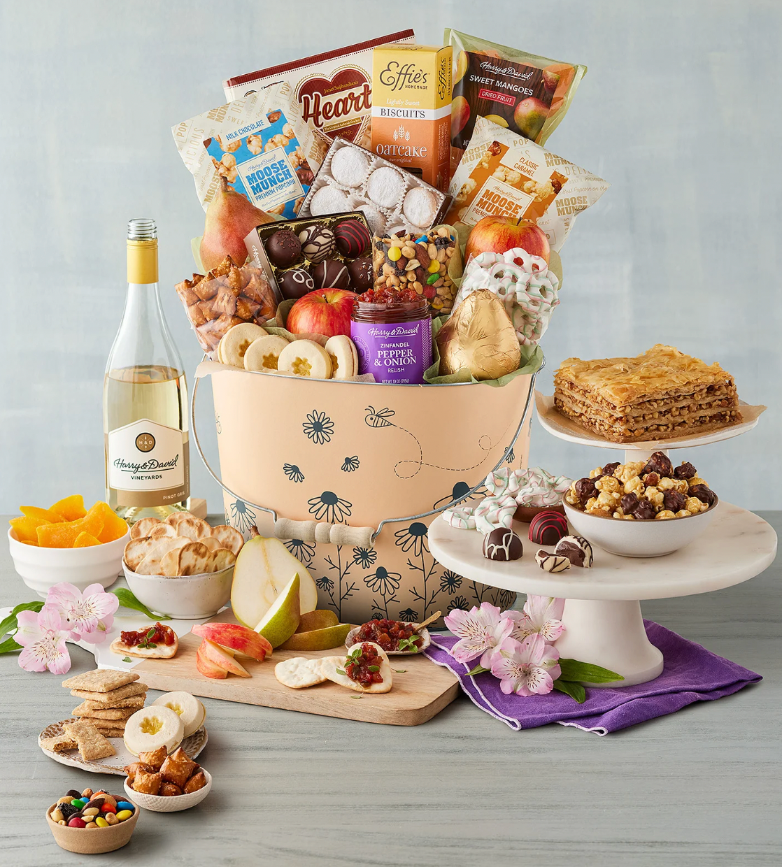 A gift basket filled with gourmet snacks, fruit, and wine, accompanied by cookies and treats on a tiered display stand.