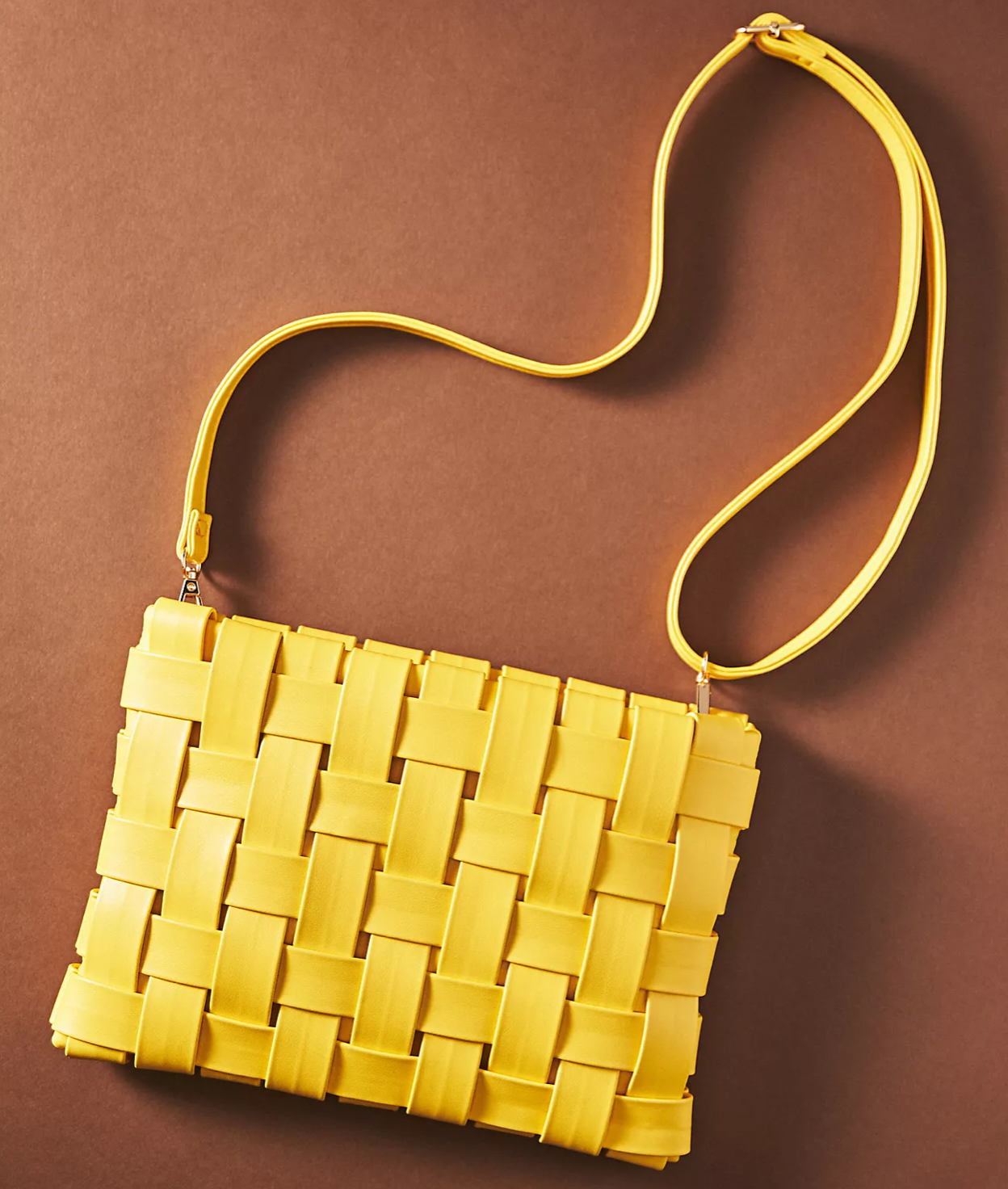 A bright yellow, woven-style crossbody bag with a slim shoulder strap, centered against a solid brown background.