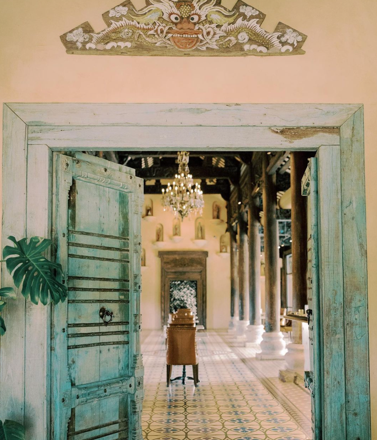 An open, teal-painted wooden door reveals a long, elegant hall with tiled floors, wooden pillars, and a crystal chandelier.