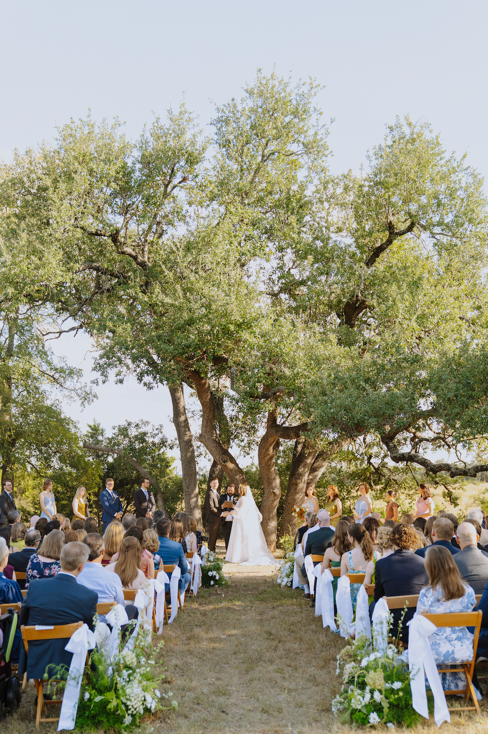 A bride and groom stand before a large, leafy tree during an outdoor wedding ceremony with guests seated in rows.