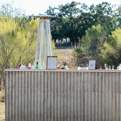 An outdoor bar with a an outdoor metal front sits in a grassy area, featuring a tall patio heater and assorted bottles. 