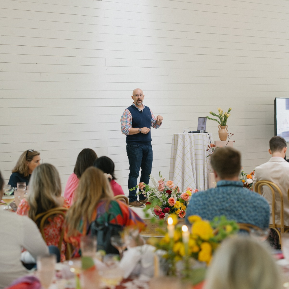 Speaker addressing seated audience in a bright event hall, with floral centerpieces on the tables.