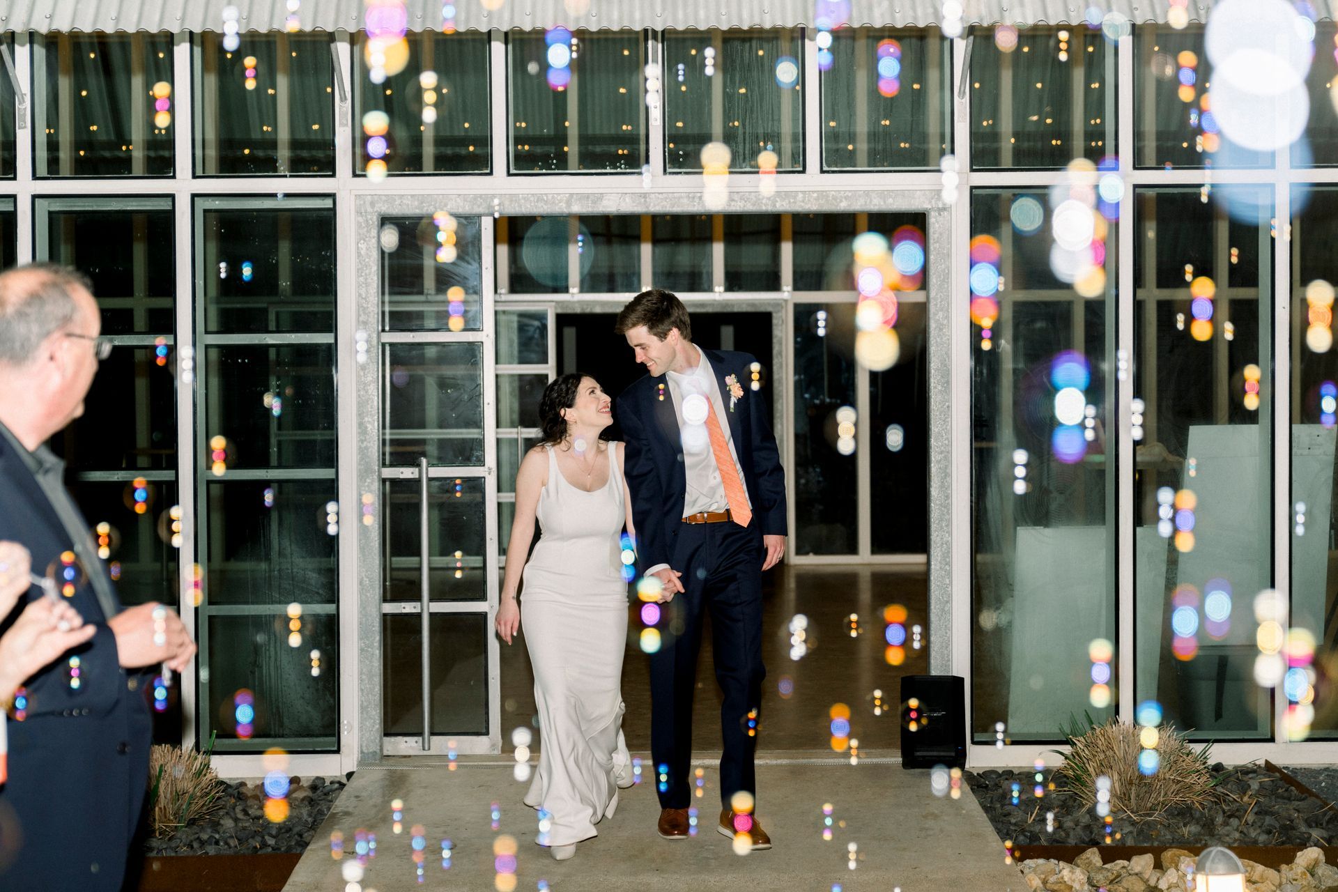 A couple smiles while walking through a doorway, surrounded by floating bubbles, outside a glass-walled building at night at Prospect House. 
