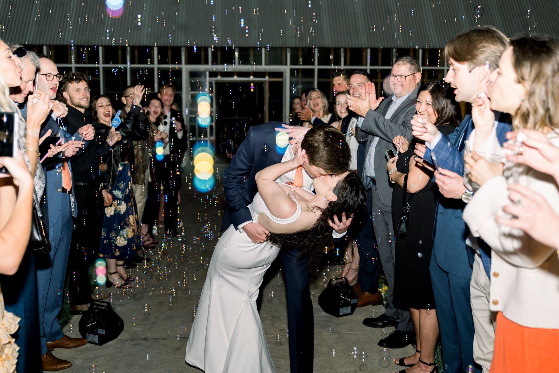 A couple in wedding attire shares a dip and kiss amid floating bubbles, surrounded by cheering guests at a celebration.