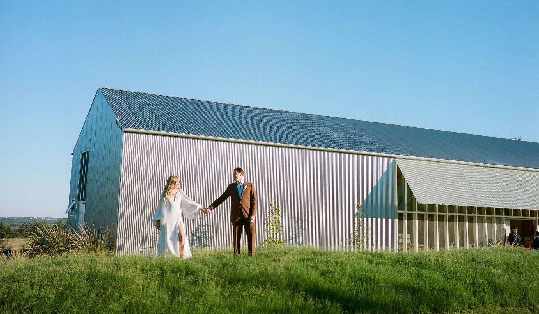 A couple holding hands walks through a grassy field in front of a modern corrugated metal barn under a clear blue sky.