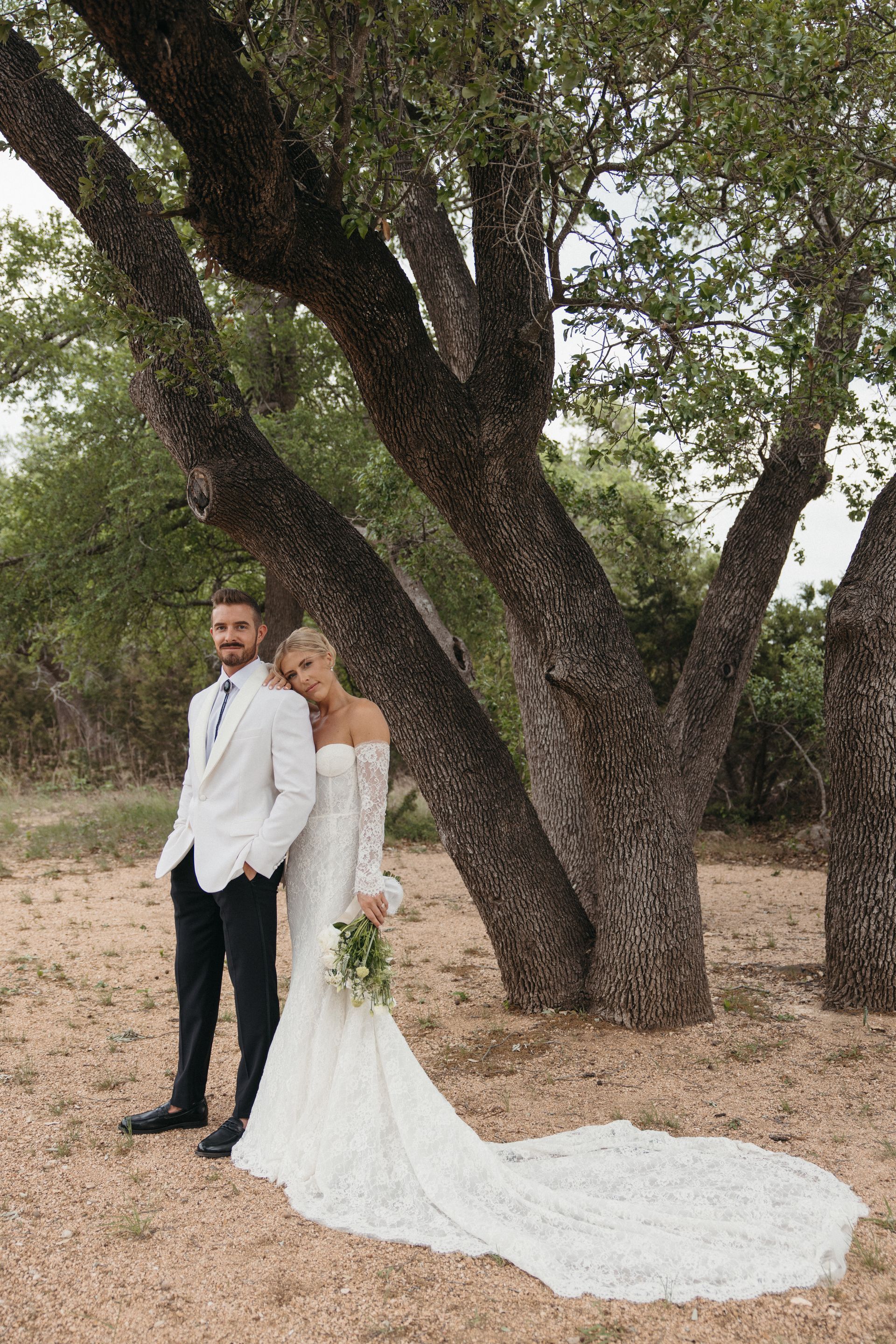 A couple in wedding attire poses together under a large tree in a natural, outdoor setting.