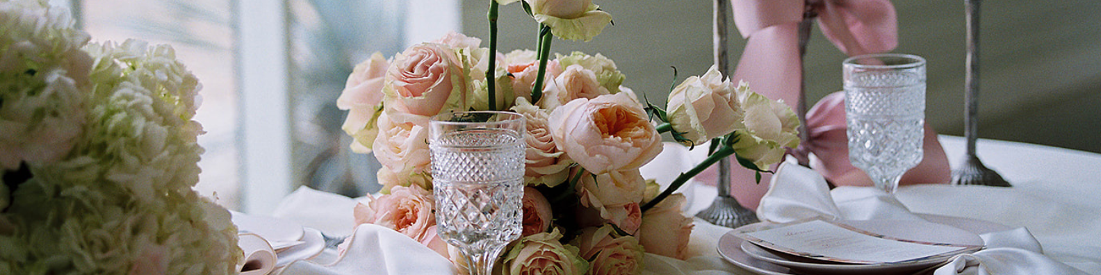 A table set for a formal event with pale pink rose centerpieces, crystal glasses, and white linens in a bright room.