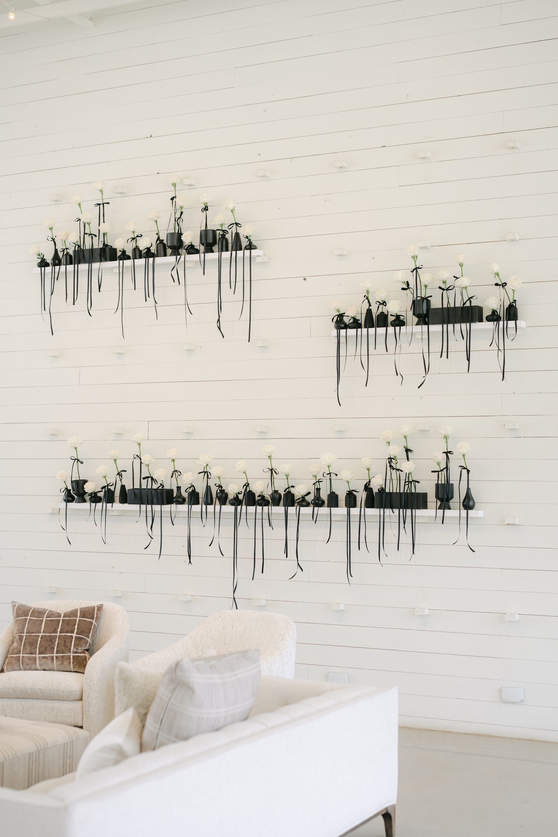 Three black shelves with white flowers and hanging black ribbons are displayed on a white brick wall above a white sofa at Prospect House.