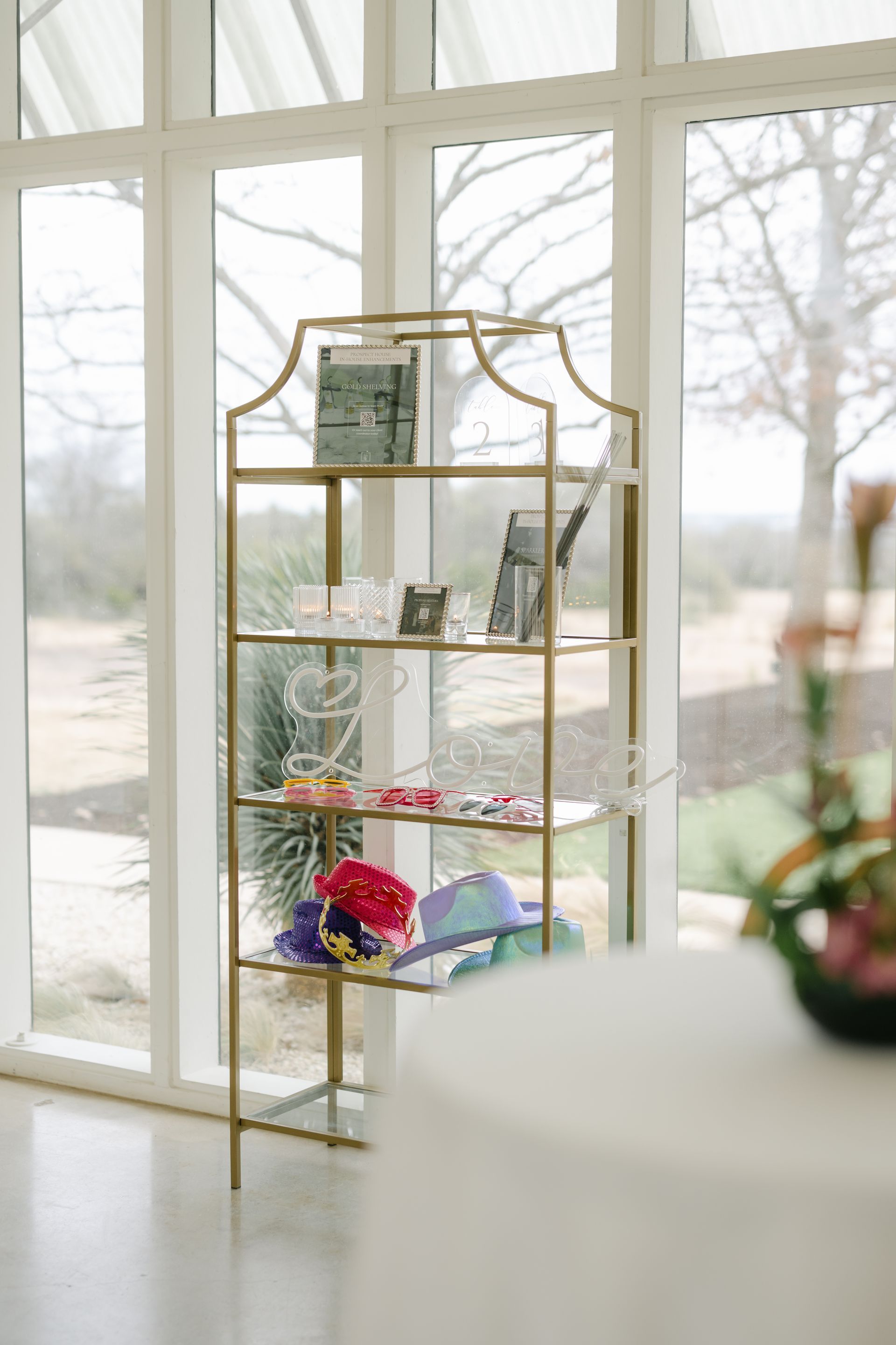 A gold-framed display shelf with multiple levels stands in a bright room, holding various accessories and items.