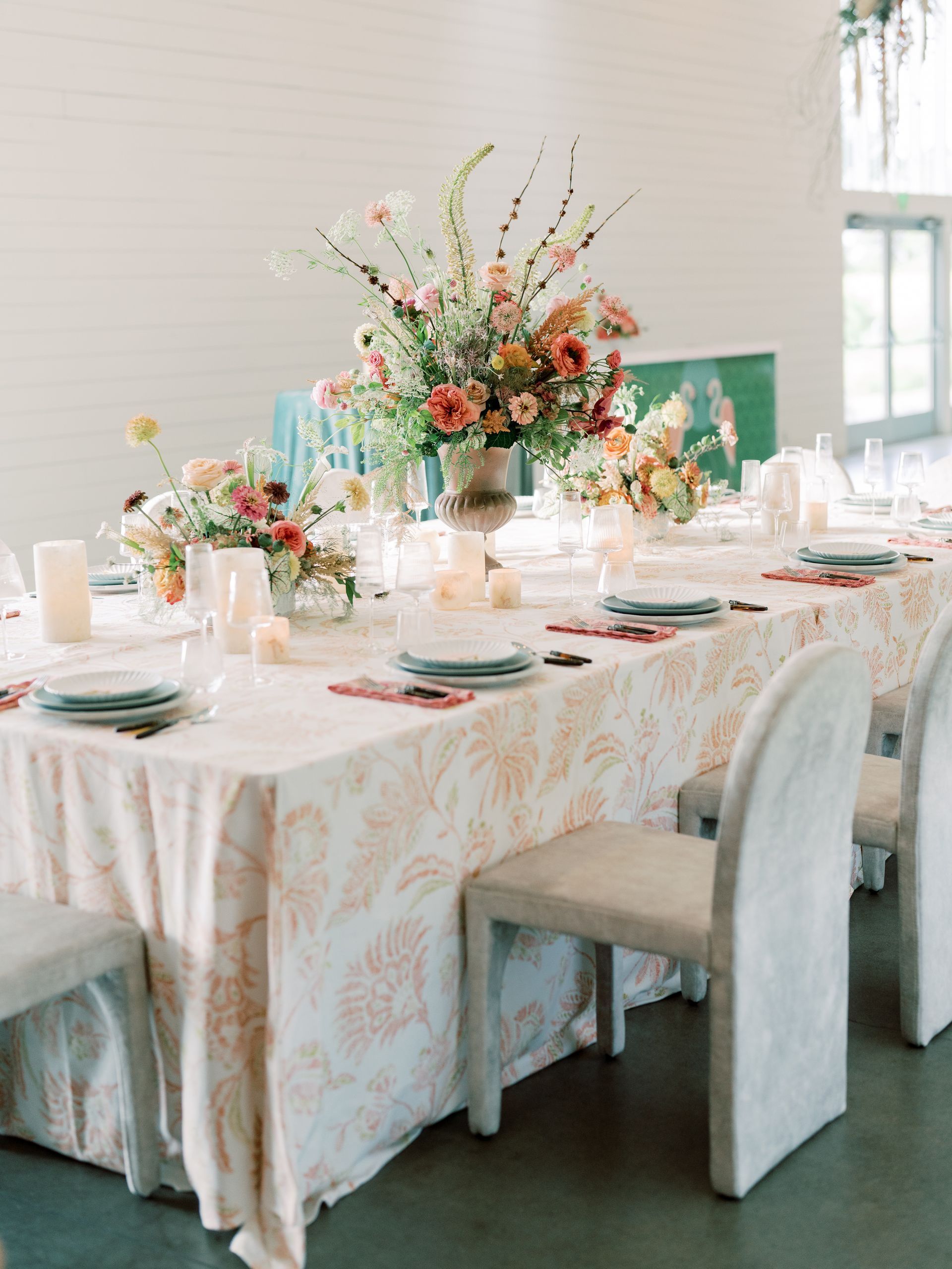A long table with a white tablecloth, and garden flowers in shades of red and pink on it.