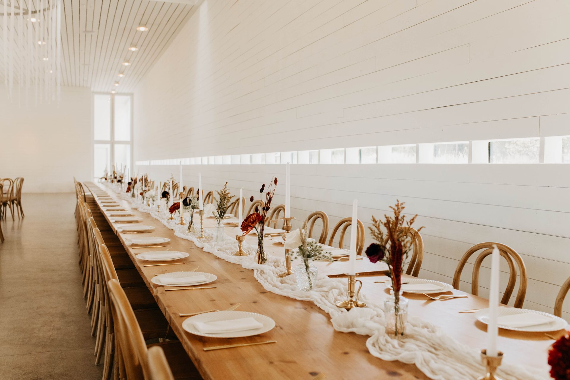 A long wooden table in a plain white room with plates, small flower votives and candles on it.