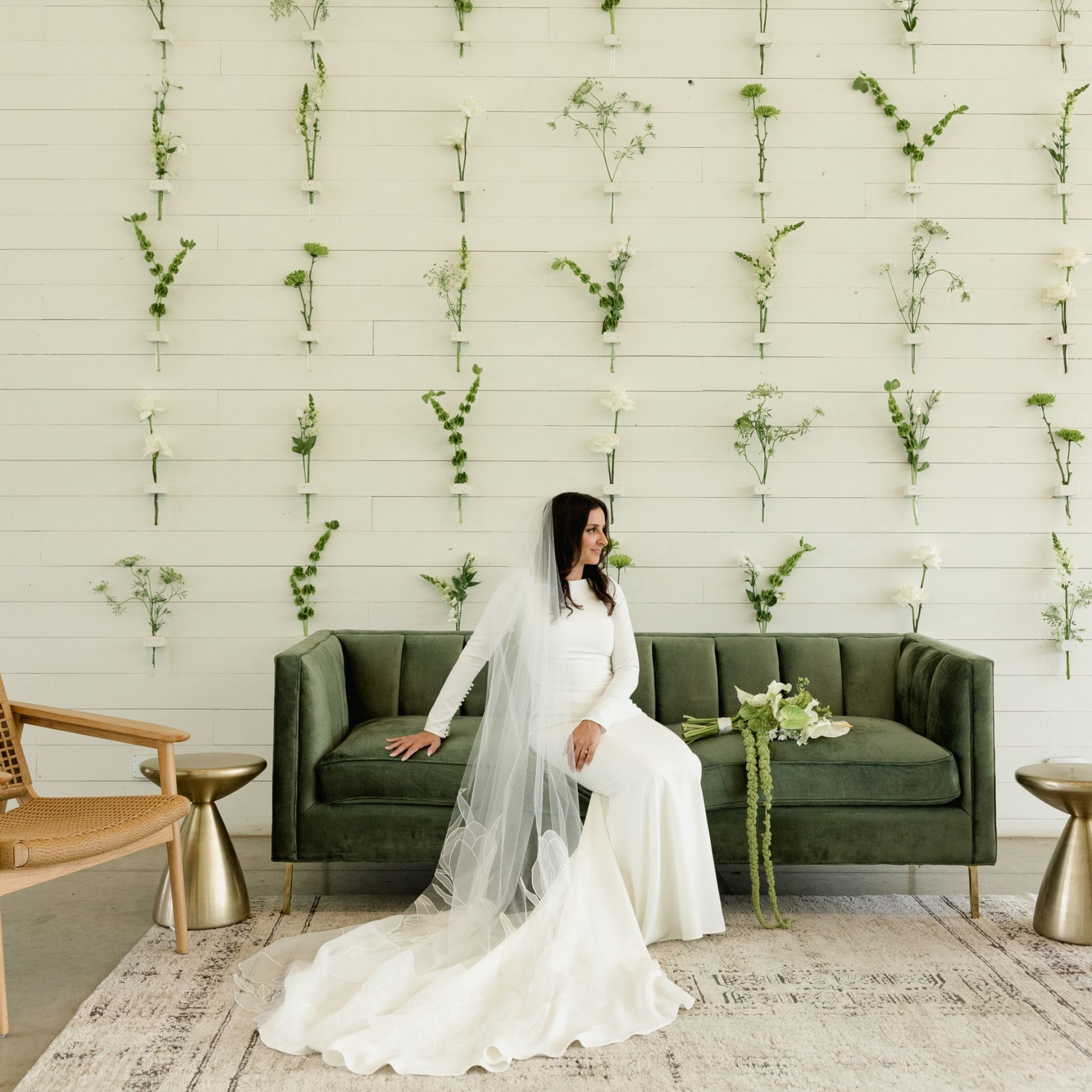 A bride in a long-sleeved white gown and veil sits on a green velvet sofa against a wall decorated with floral stems.