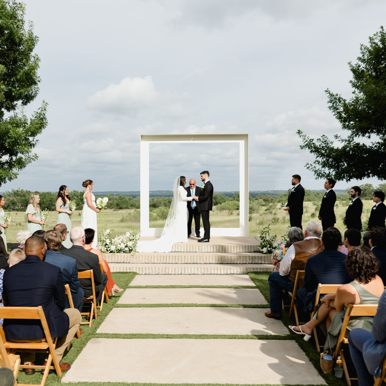 A couple exchanges vows at an outdoor wedding ceremony framed by a large white arch overlooking a rural landscape.
