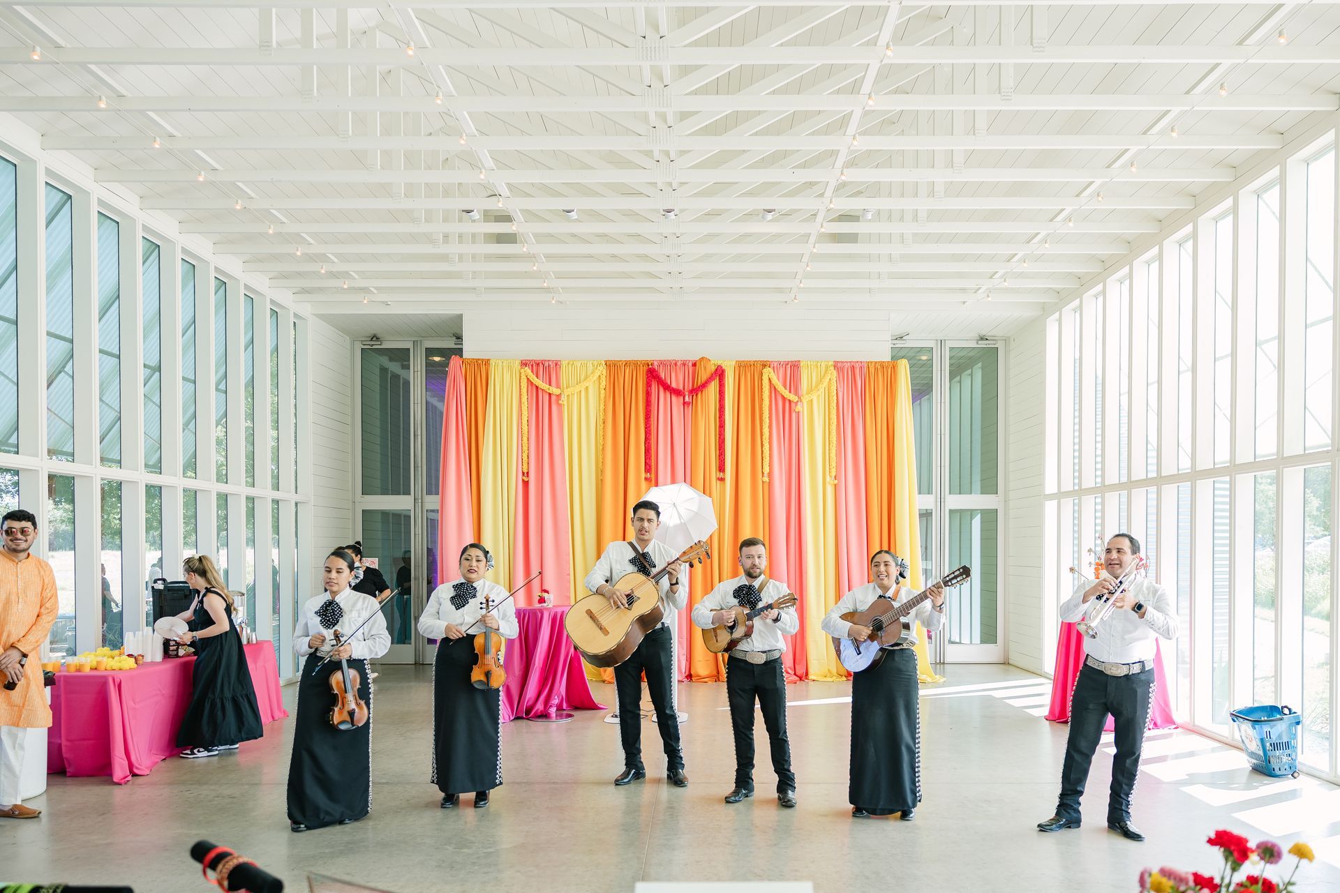 A mariachi band performs in a bright, modern hall decorated with orange, yellow, and pink curtains.