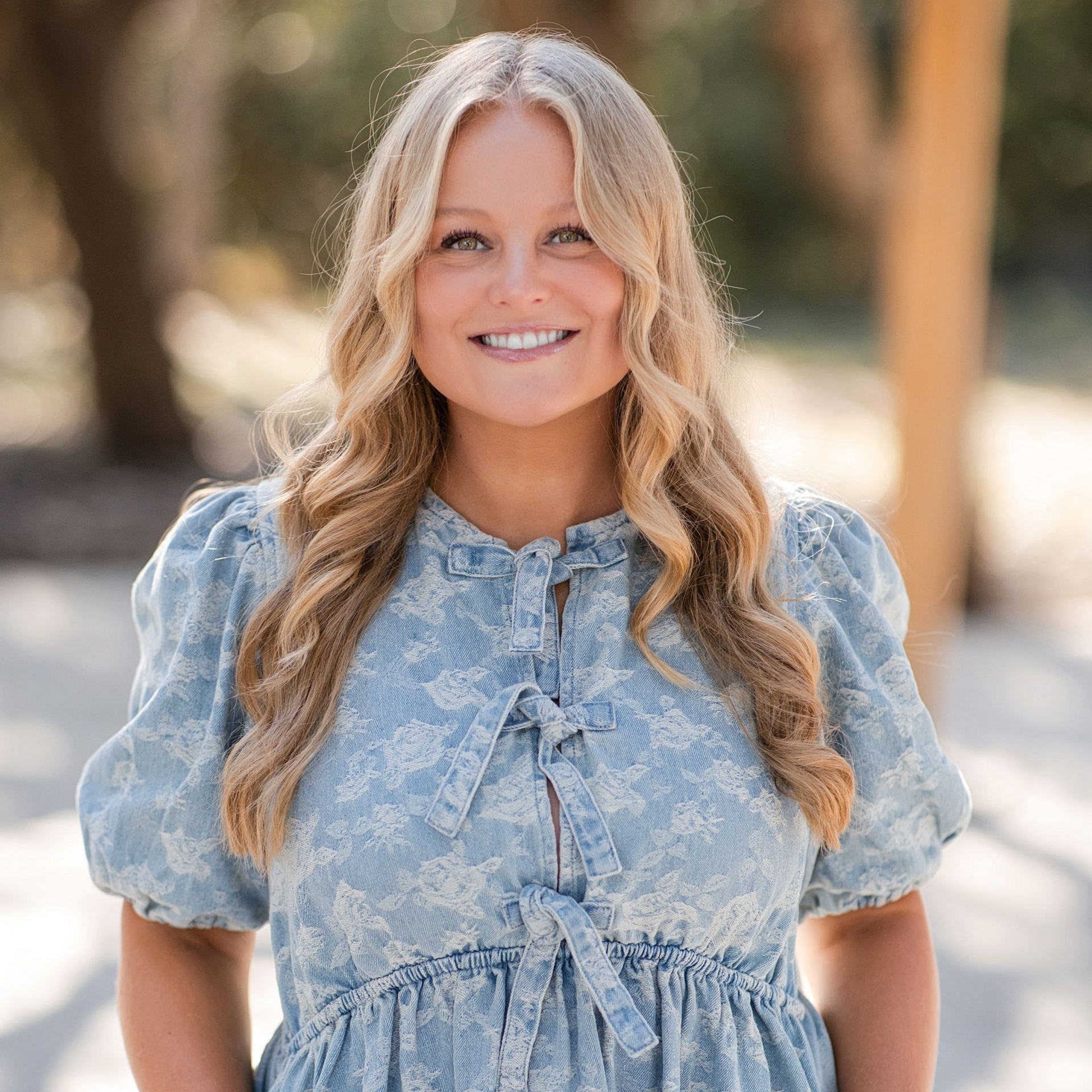 Smiling person with long blonde hair wearing a light blue, patterned puff-sleeve dress outdoors in soft lighting.