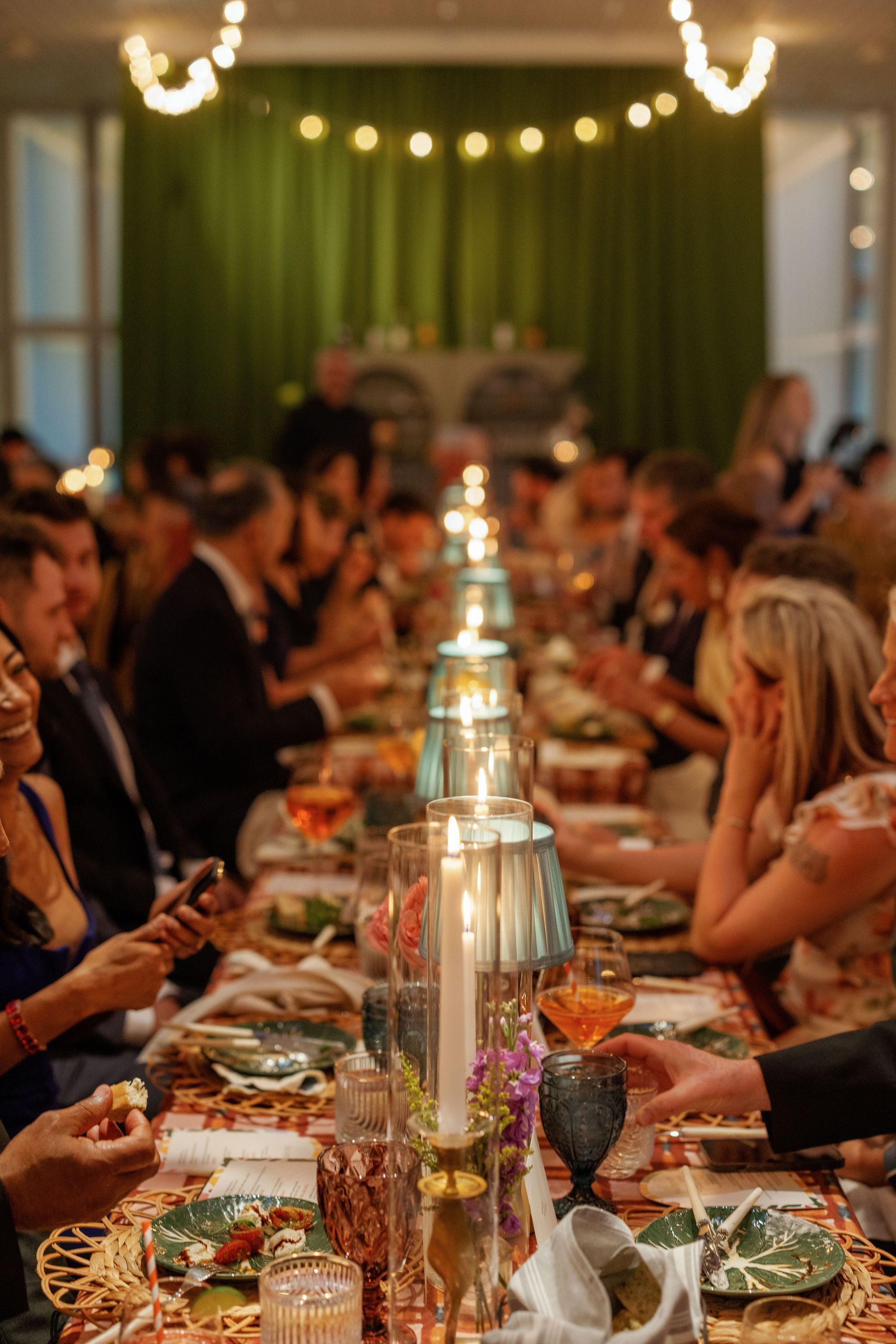 A long dinner table set with candles and tableware at a festive event, with guests gathered and engaged in conversation.