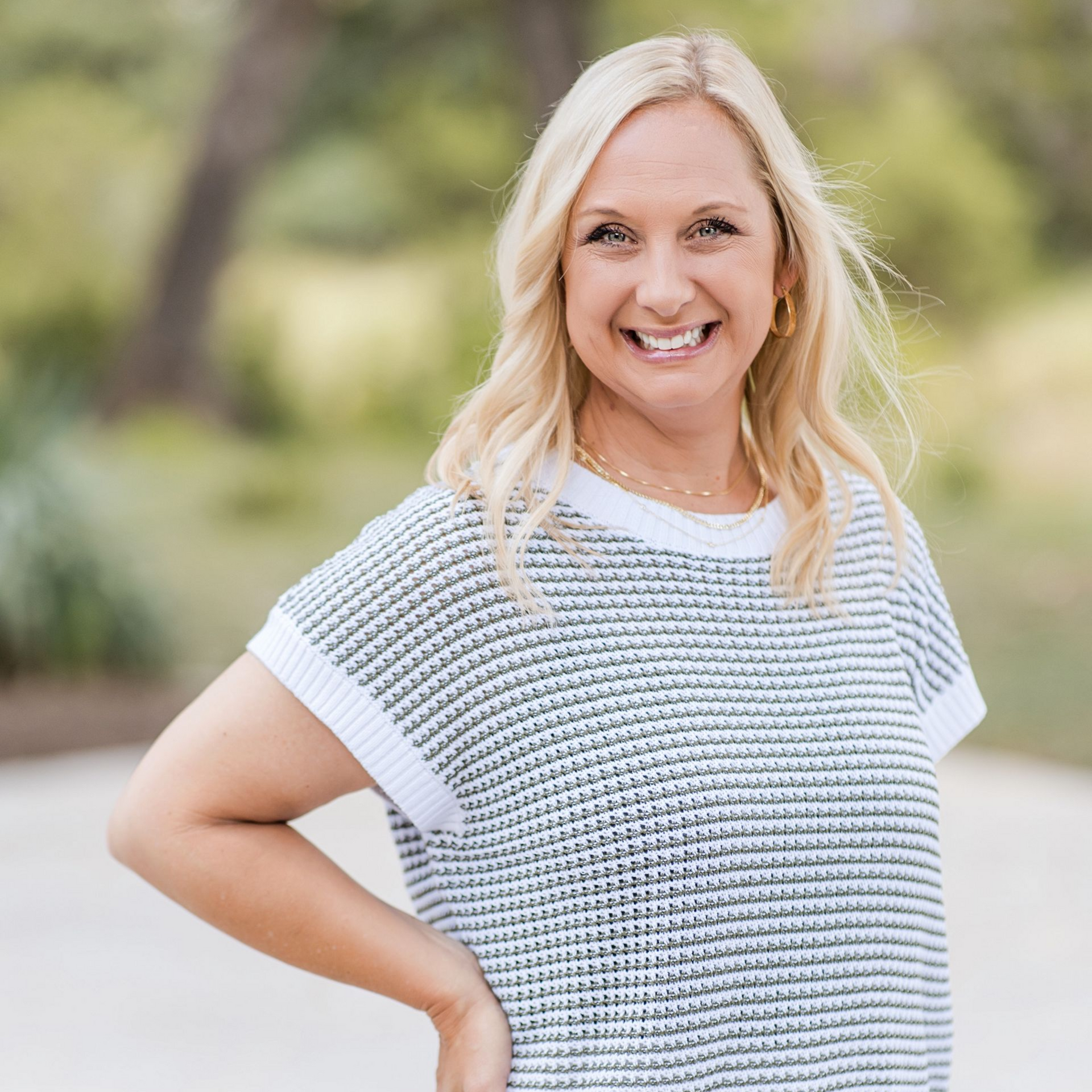 A smiling woman with blonde hair, wearing a white and grey textured short-sleeved sweater, stands outdoors.
