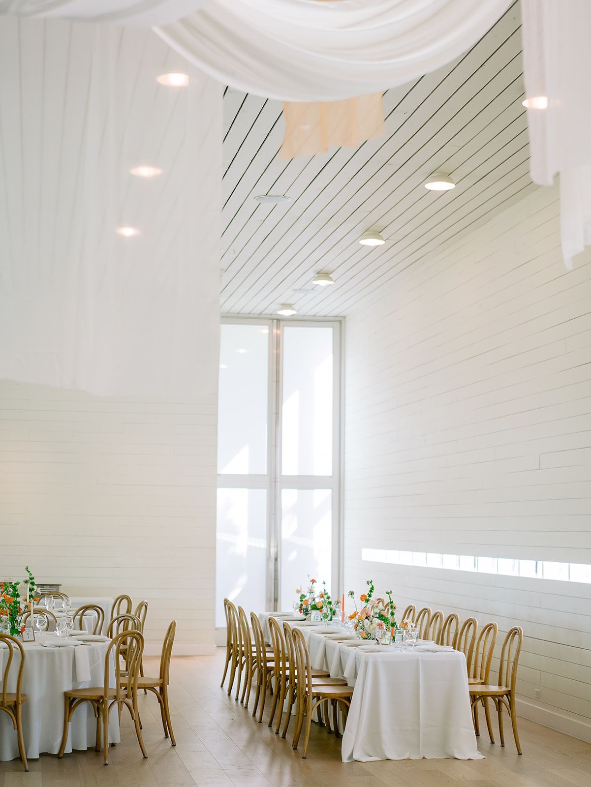 A bright, airy event space with white walls, draped fabric ceilings, and long tables set for a meal with wooden chairs.