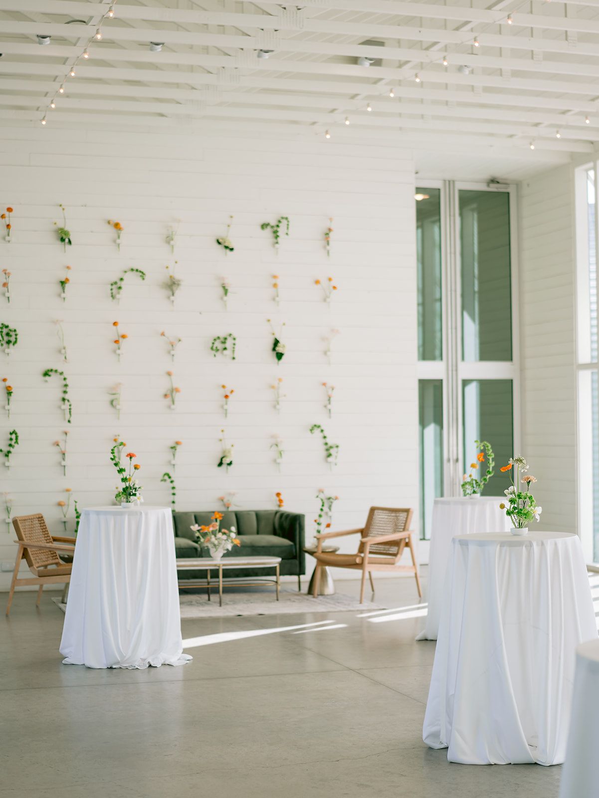 A bright event space features a wall with small floral arrangements, a green velvet sofa, and tall white-covered tables.