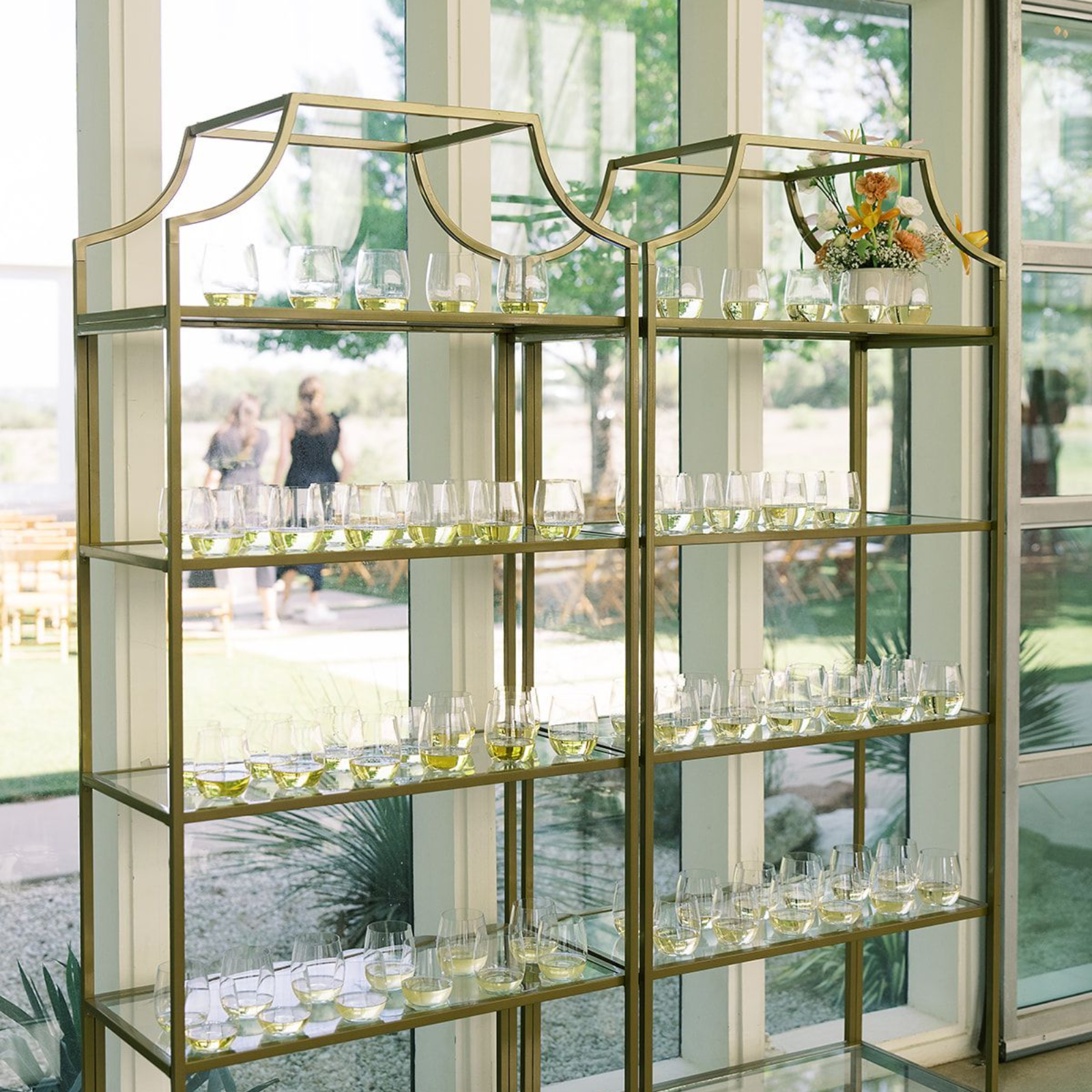 Two tall, gold, tiered metal shelves filled with rows of clear glassware, positioned in front of a bright window at Prospect House.